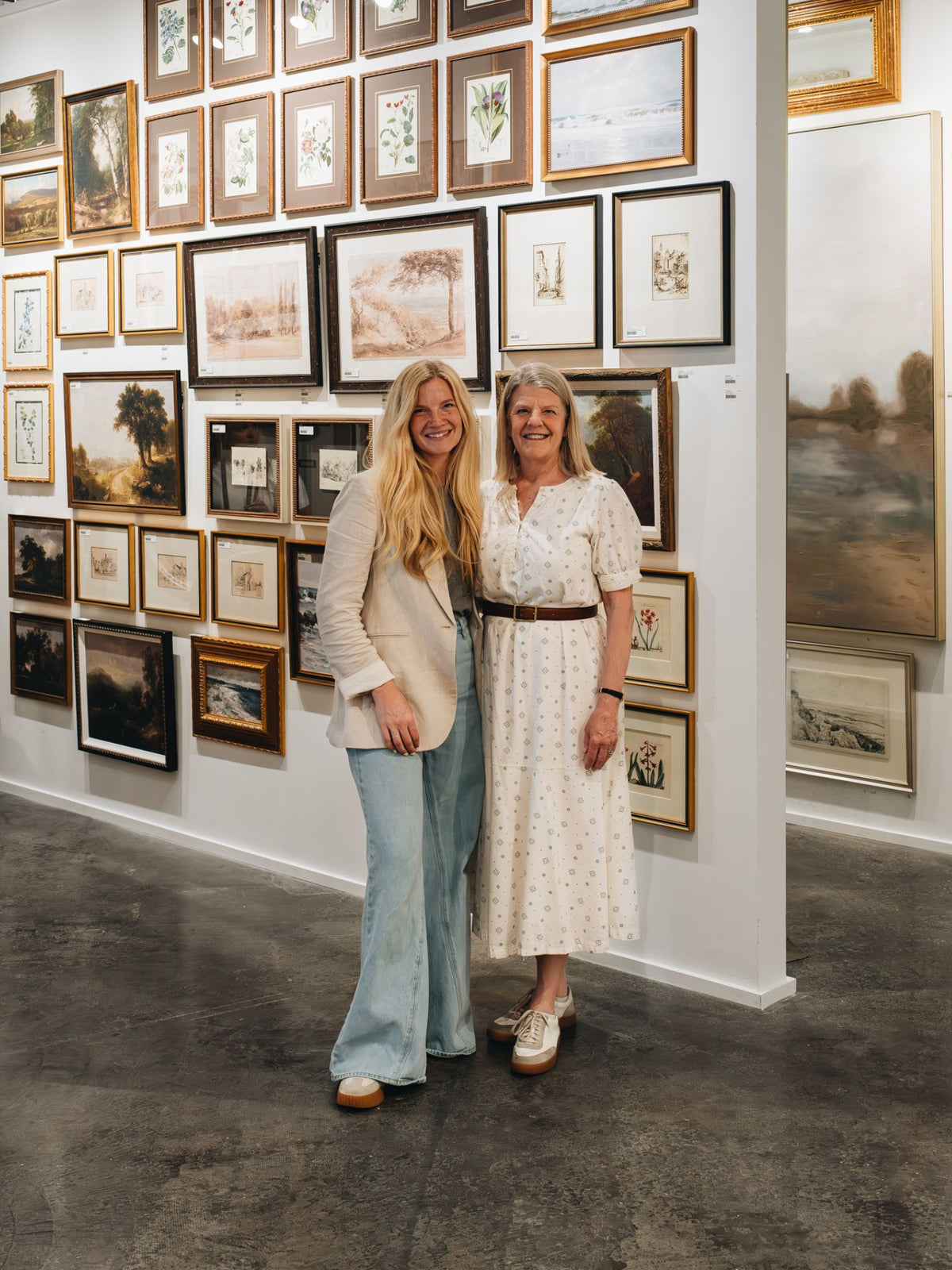 Two women standing together in an art gallery, smiling in front of a wall covered with framed landscape and botanical prints. One wears a cream blazer and light blue jeans, and the other wears a white patterned dress with a brown belt.