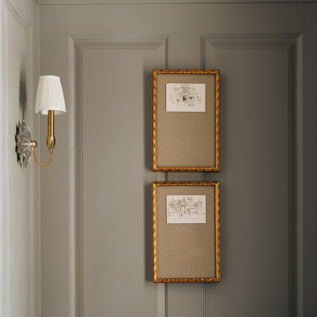 Two vertical framed art prints with gold rope-textured frames hang on a gray paneled wall next to a brass and white wall sconce.