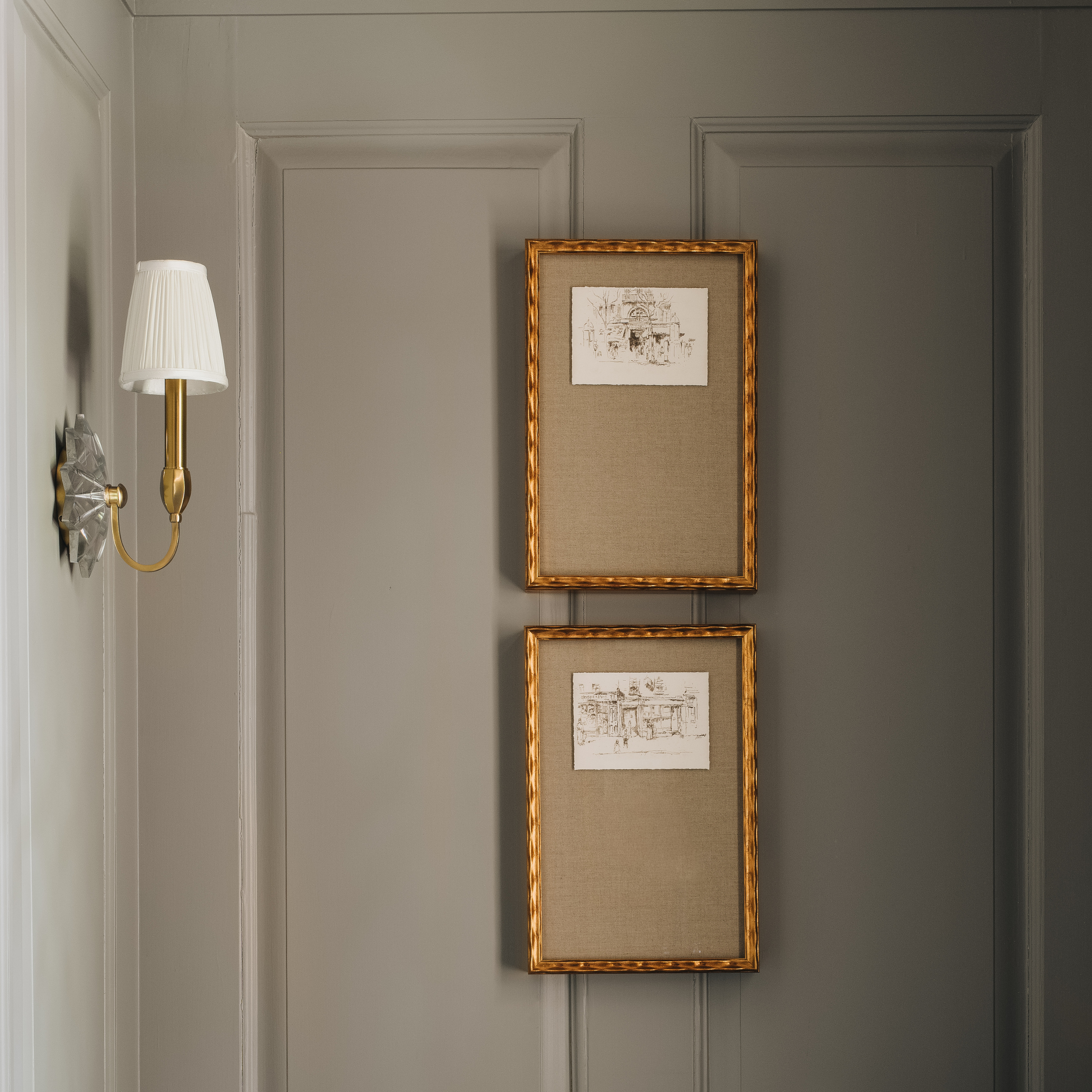 Two vertical framed art prints with gold rope-textured frames hang on a gray paneled wall next to a brass and white wall sconce.