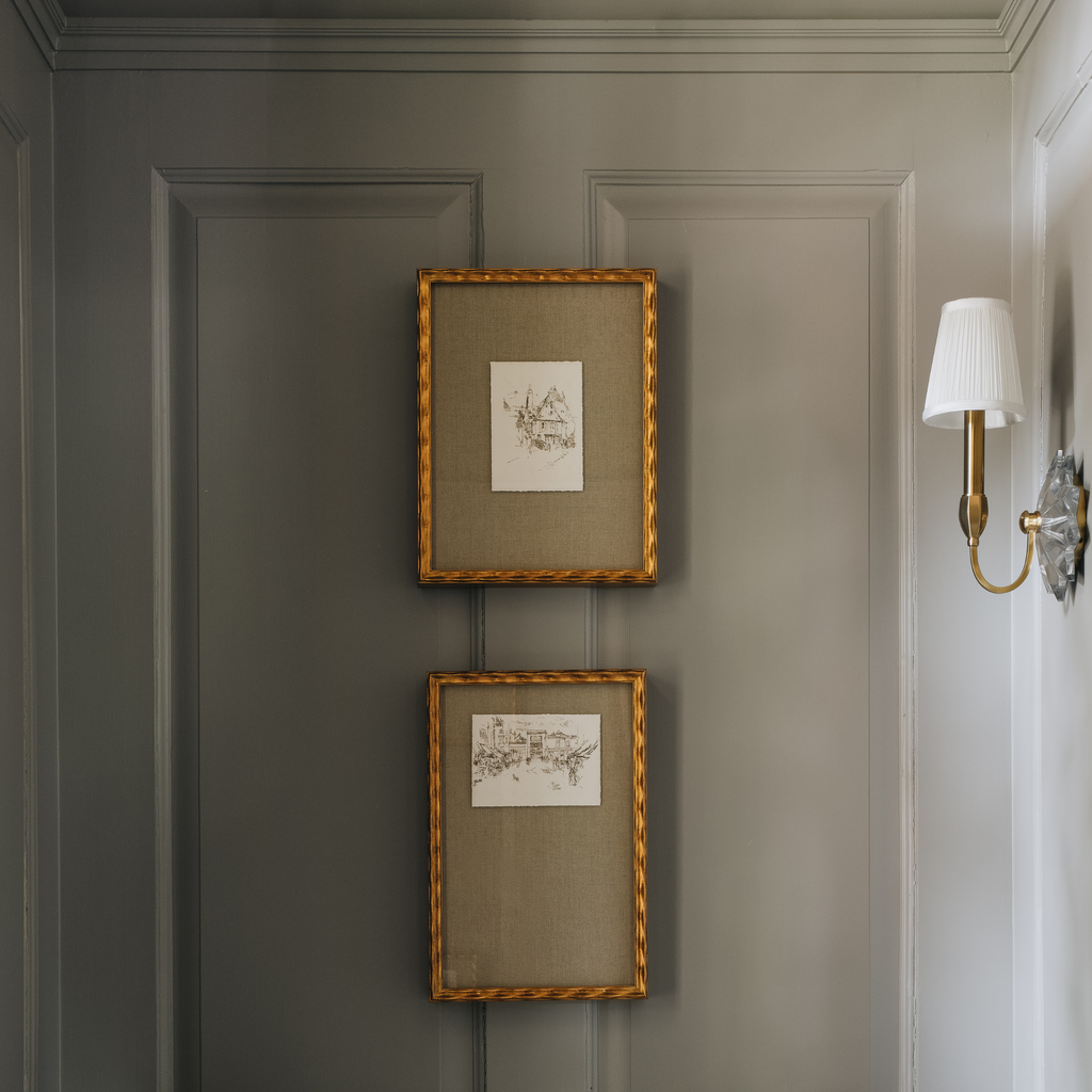 Two small framed botanical art prints with gold frames hang vertically on a gray paneled wall next to a white wall sconce.