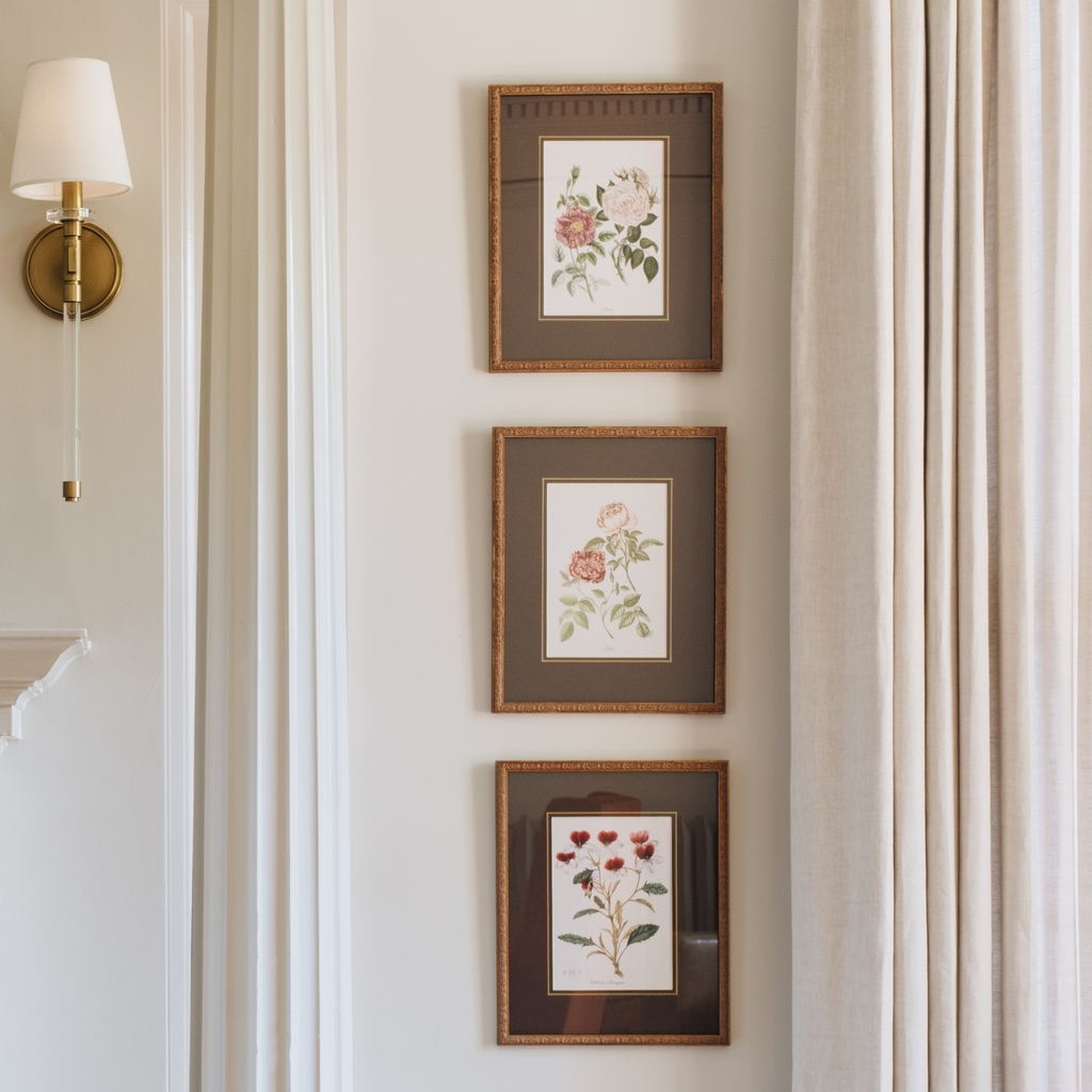 Three framed botanical art prints with pink and red flowers are arranged vertically on a white wall next to a gold wall sconce and beige curtains.