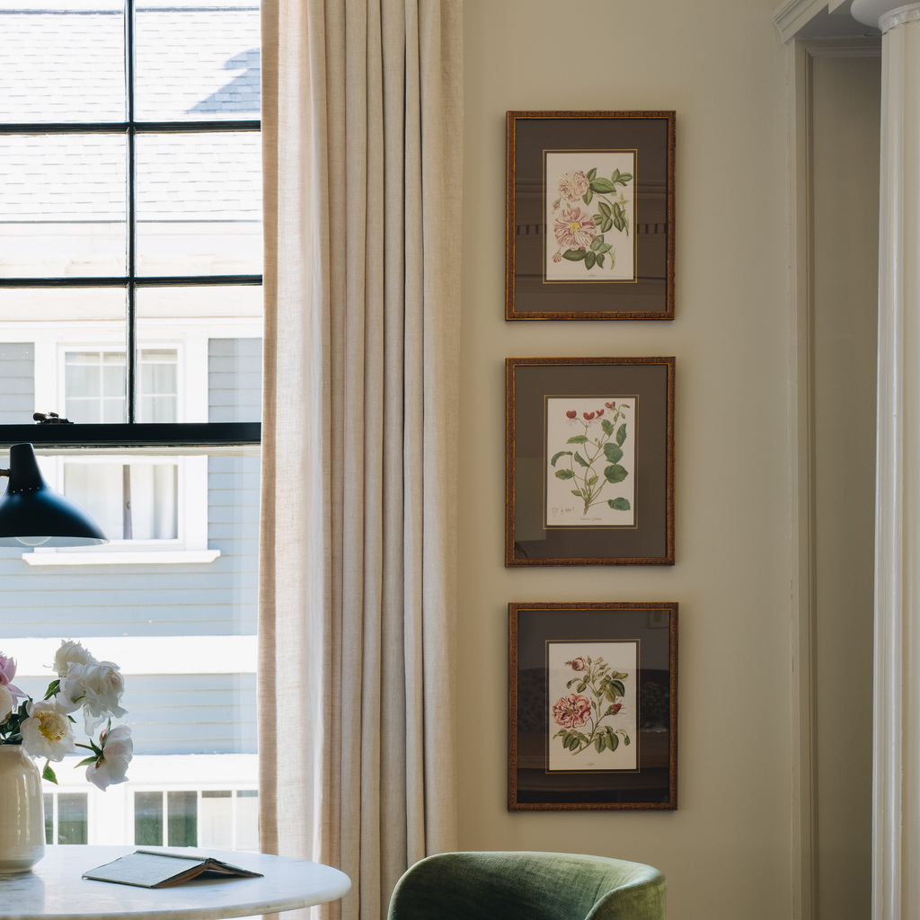 Three framed botanical art prints with pink and green flowers hang vertically on a beige wall in a bright room next to a window with sheer curtains.