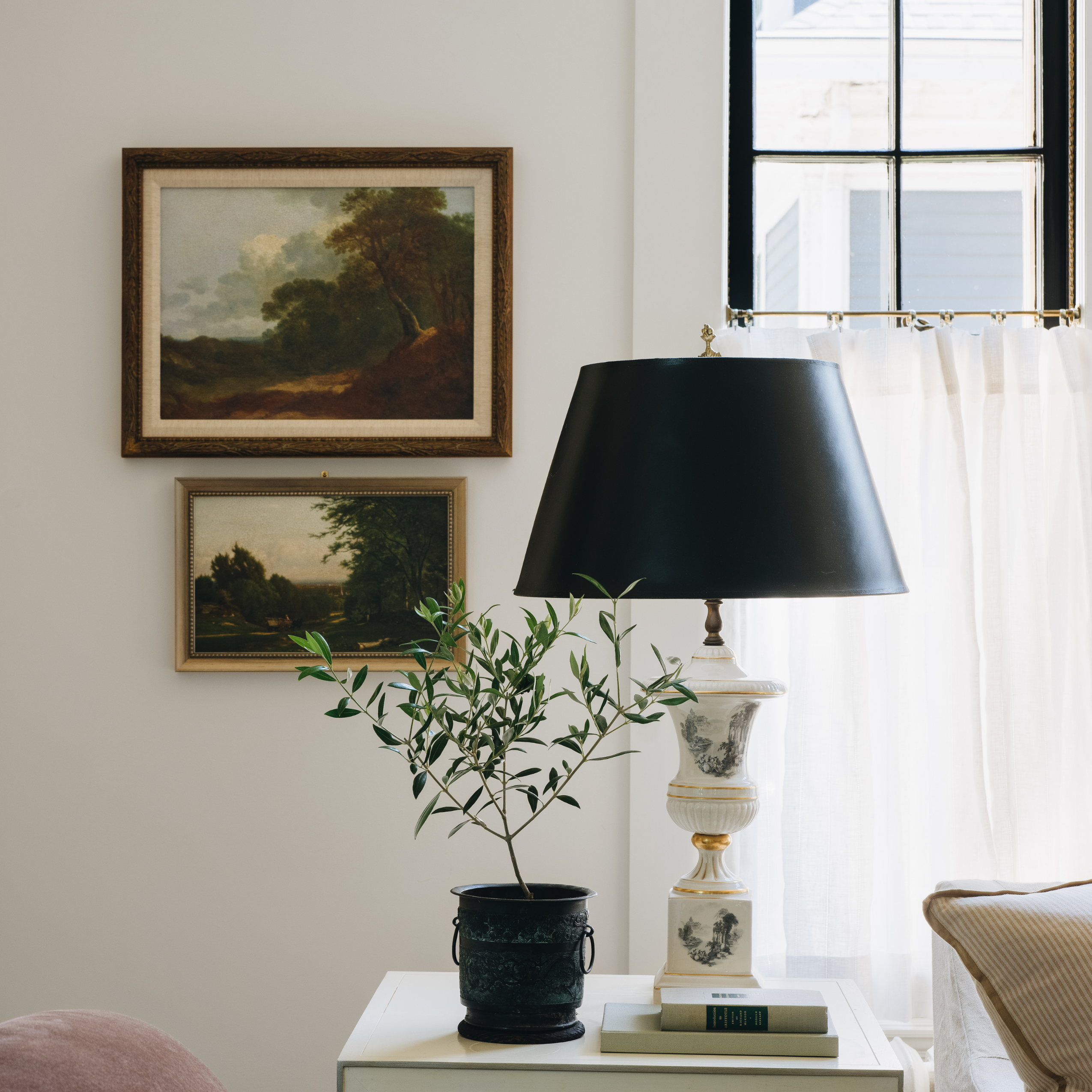 A black lampshade table lamp with a white and gold ceramic base sits on a white side table next to a potted olive tree in a dark pot. The background shows a warm, neutral-toned room with two framed landscape paintings on the wall and a window with sheer white curtains.