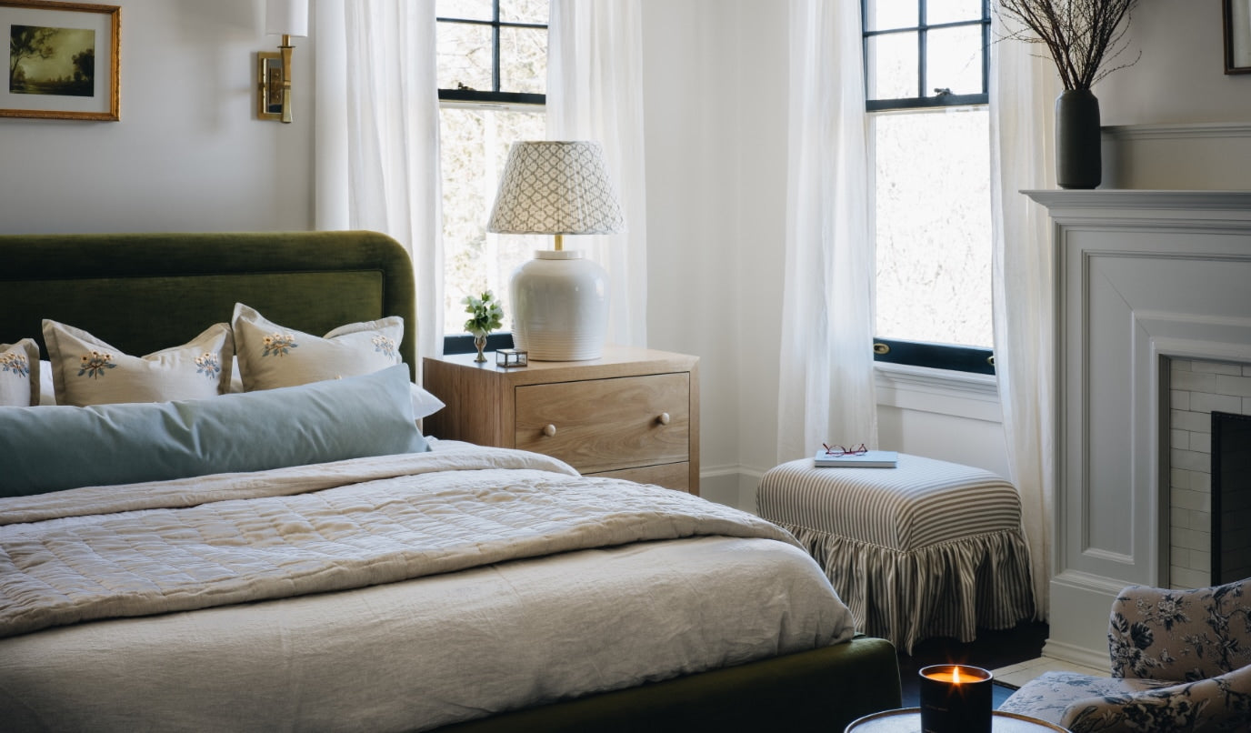 A styled bedroom interior featuring a bed with cream bedding, a green upholstered headboard, a wooden nightstand with a patterned table lamp, sheer white curtains, and a white fireplace mantel in the background.