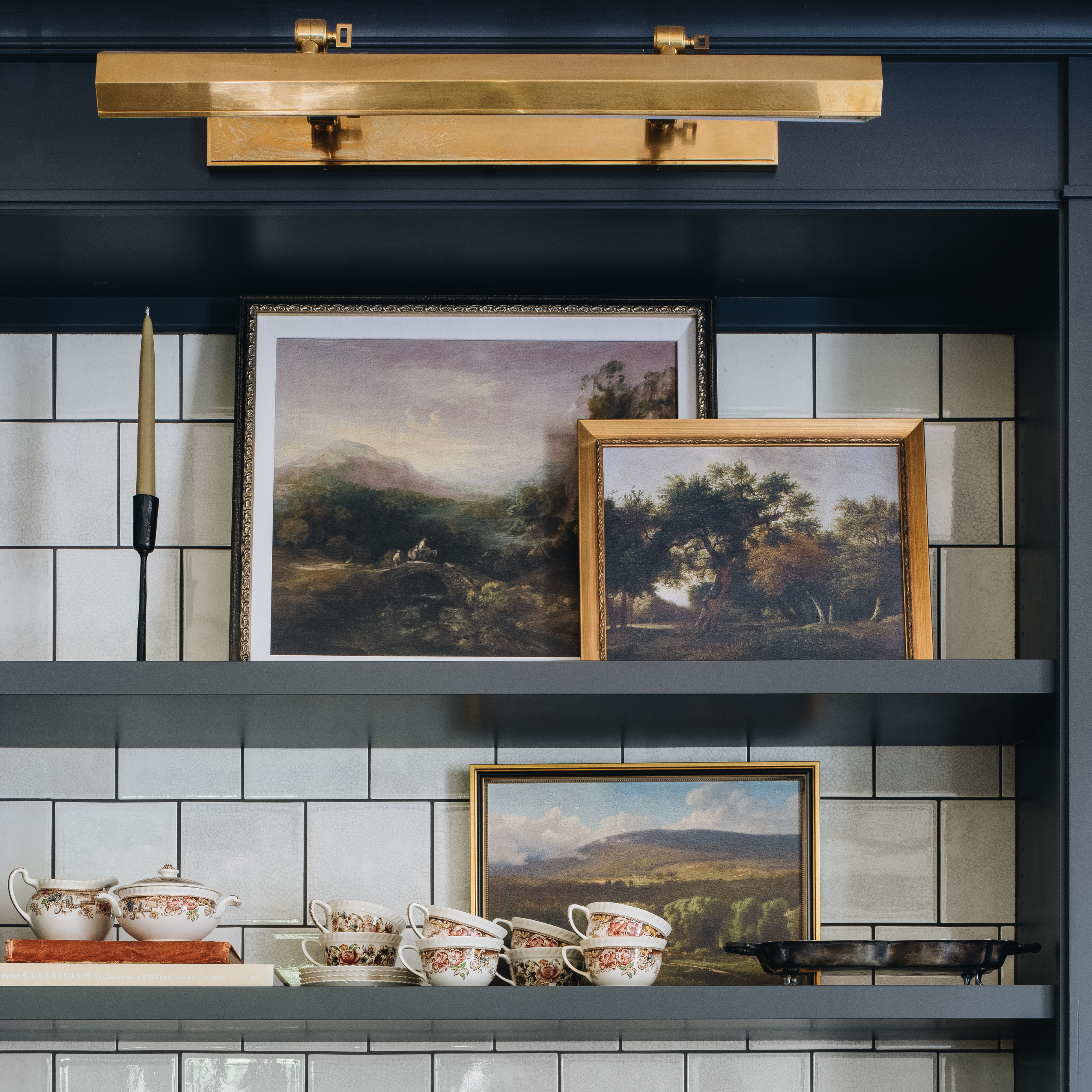 A styled interior scene with dark navy shelving, white subway tile, a brass wall light, framed landscape paintings, a candle, teacups, and a bowl.
