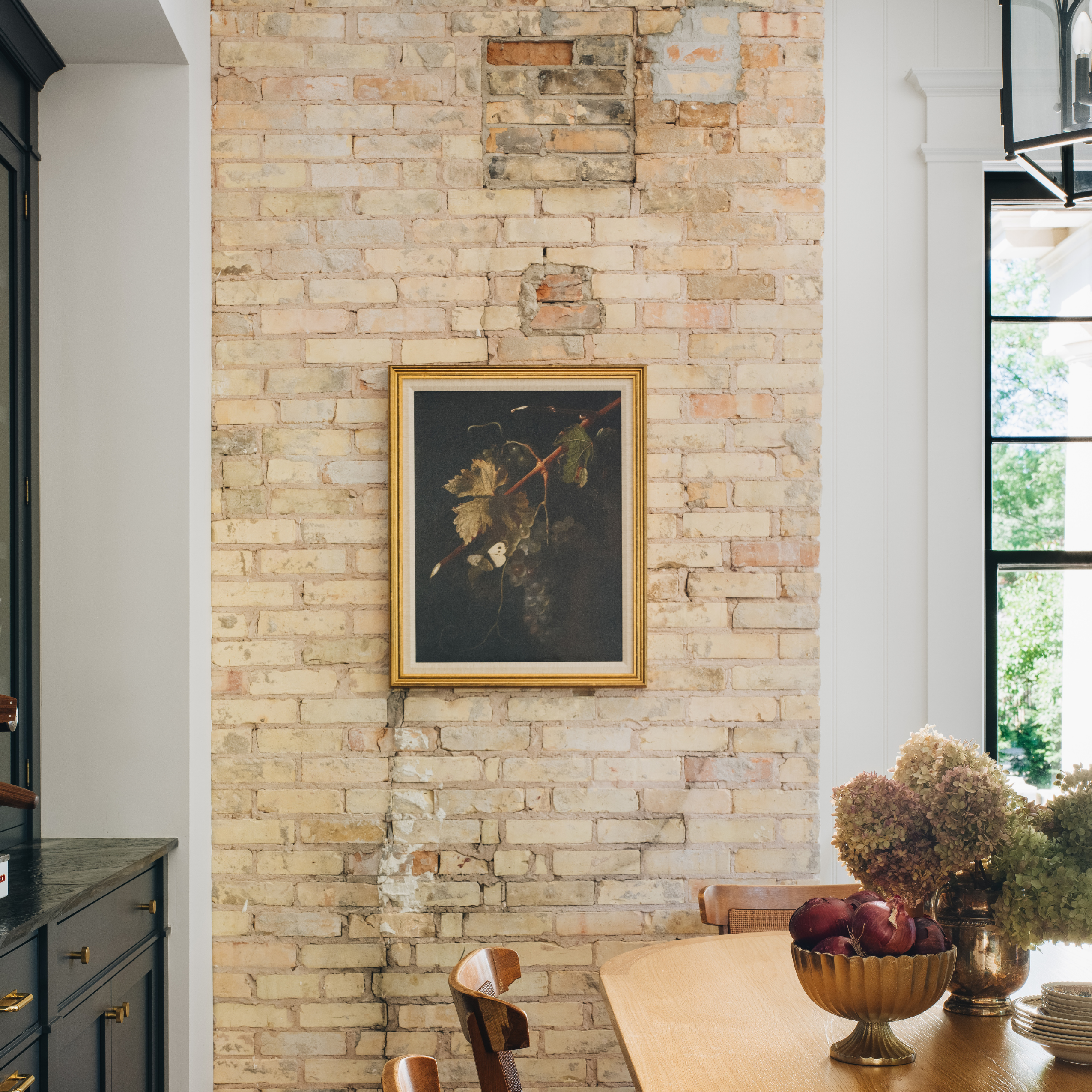 A framed still life painting with dark grapes hangs on a light brick wall in a dining room setting with a wooden table and decorative bowl of red apples.