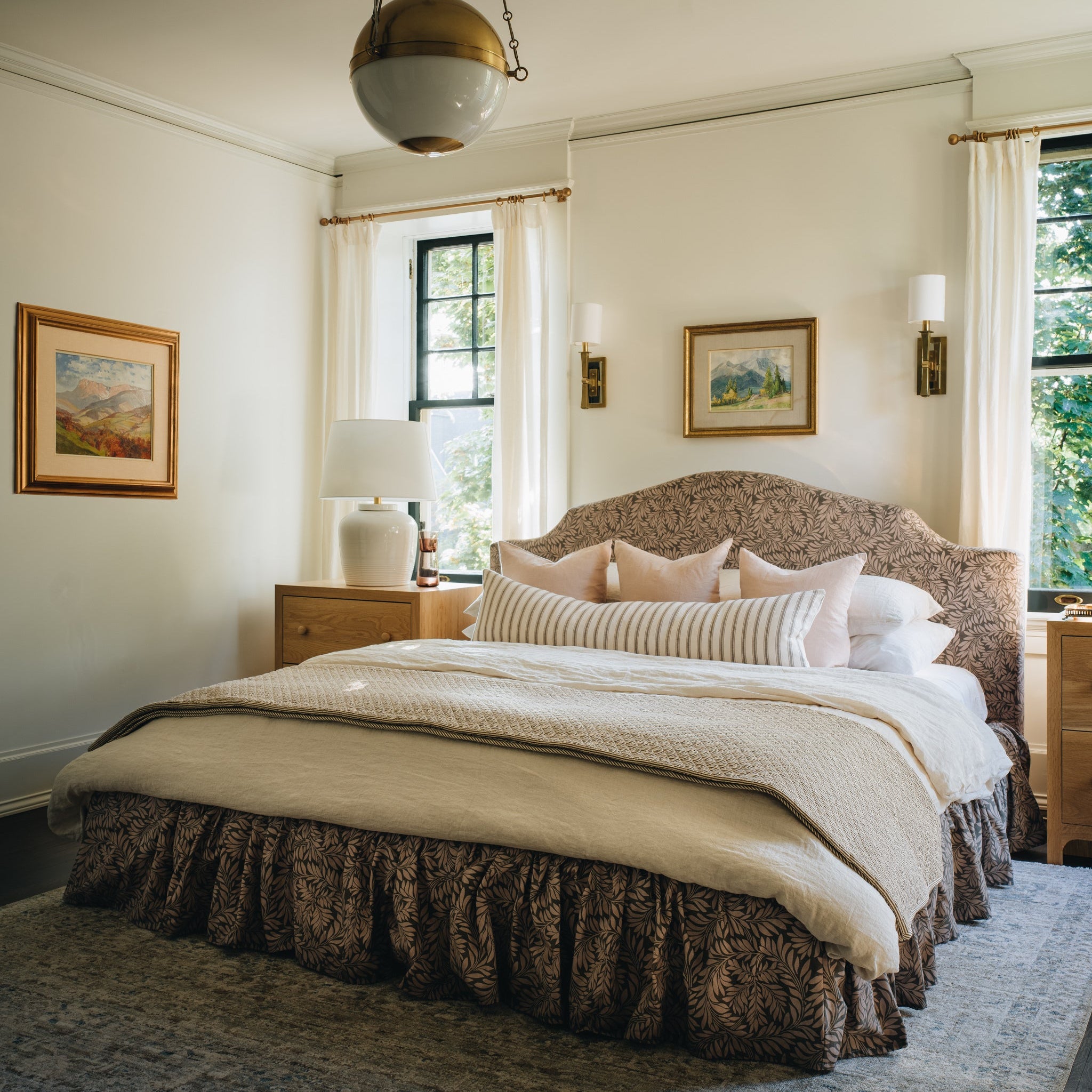 A beautifully styled bedroom featuring a large bed with a textured upholstered headboard, cream and beige bedding, a patterned brown ruffled bed skirt, and a gray patterned area rug. The room has warm off-white walls, a hanging brass and white globe pendant light, wall sconces, and framed landscape art.