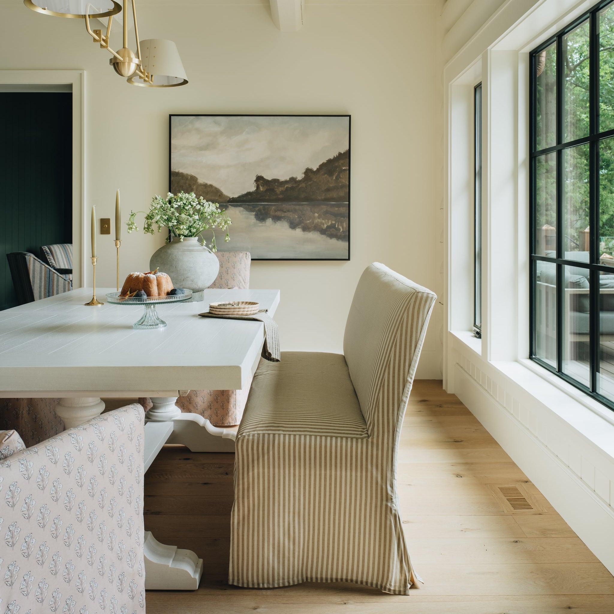 A styled dining room corner featuring a white dining table, a beige striped upholstered chair, a hanging brass chandelier, and a framed landscape painting on a cream wall.