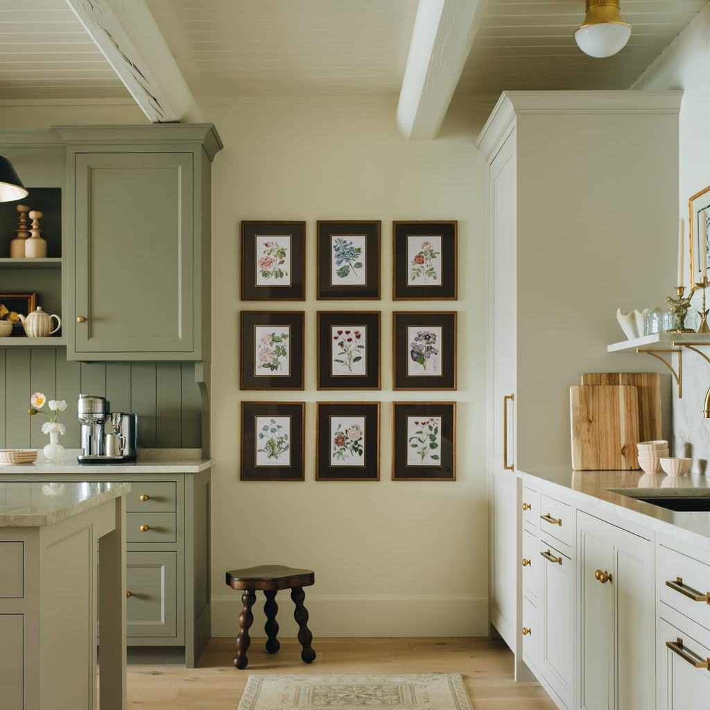 A bright, modern kitchen interior with white and sage green cabinets, a patterned runner rug, light wood floors, and brass hardware. A central wall displays a 3x3 grid of small framed botanical prints.
