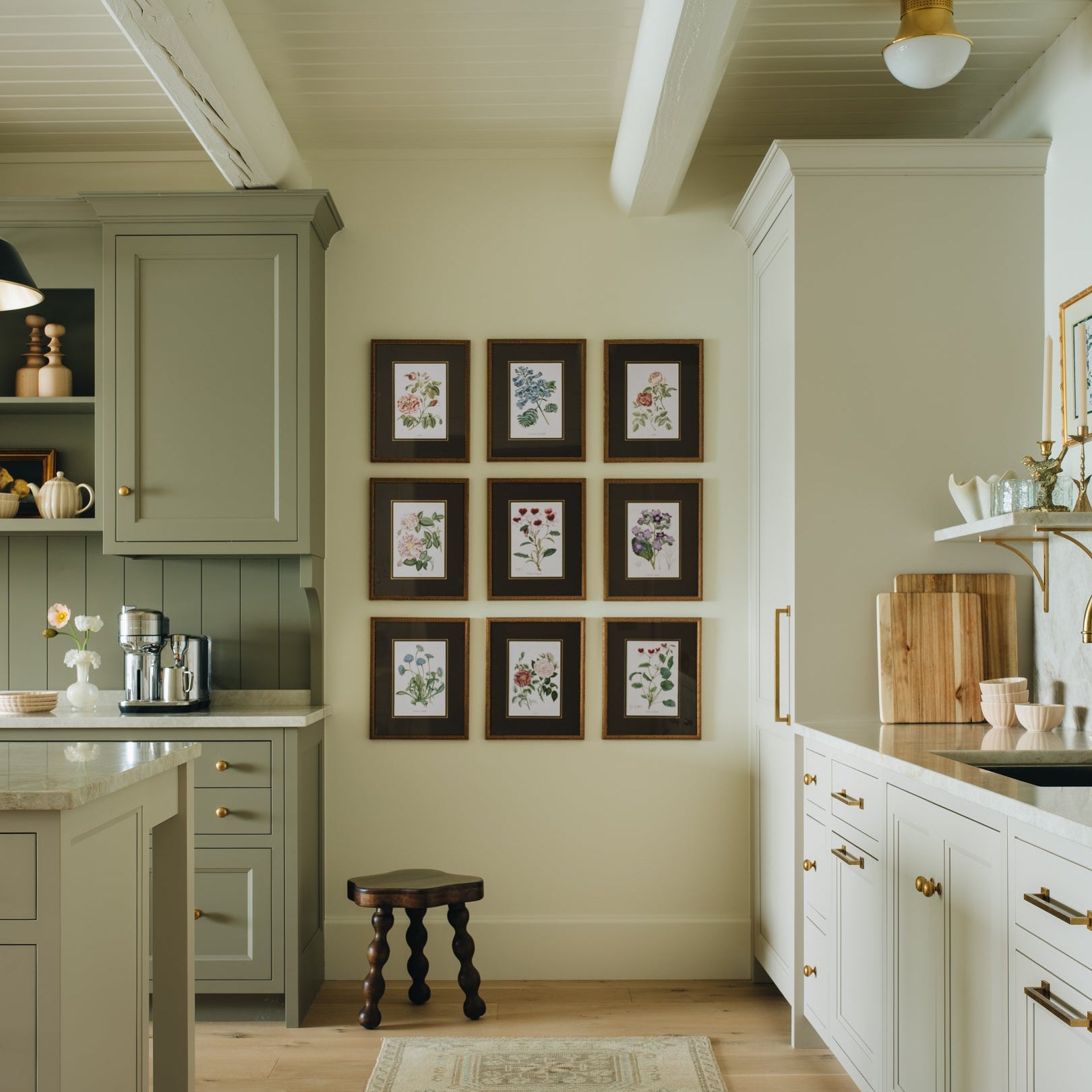 A bright, modern kitchen interior with white and sage green cabinets, a patterned runner rug, light wood floors, and brass hardware. A central wall displays a 3x3 grid of small framed botanical prints.