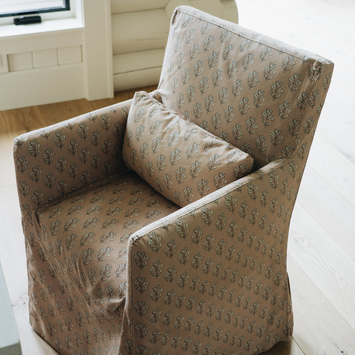A slipcovered armchair with a matching throw pillow, featuring a beige and brown leafy pattern, set in a bright, modern room with light wood floors and a log wall.