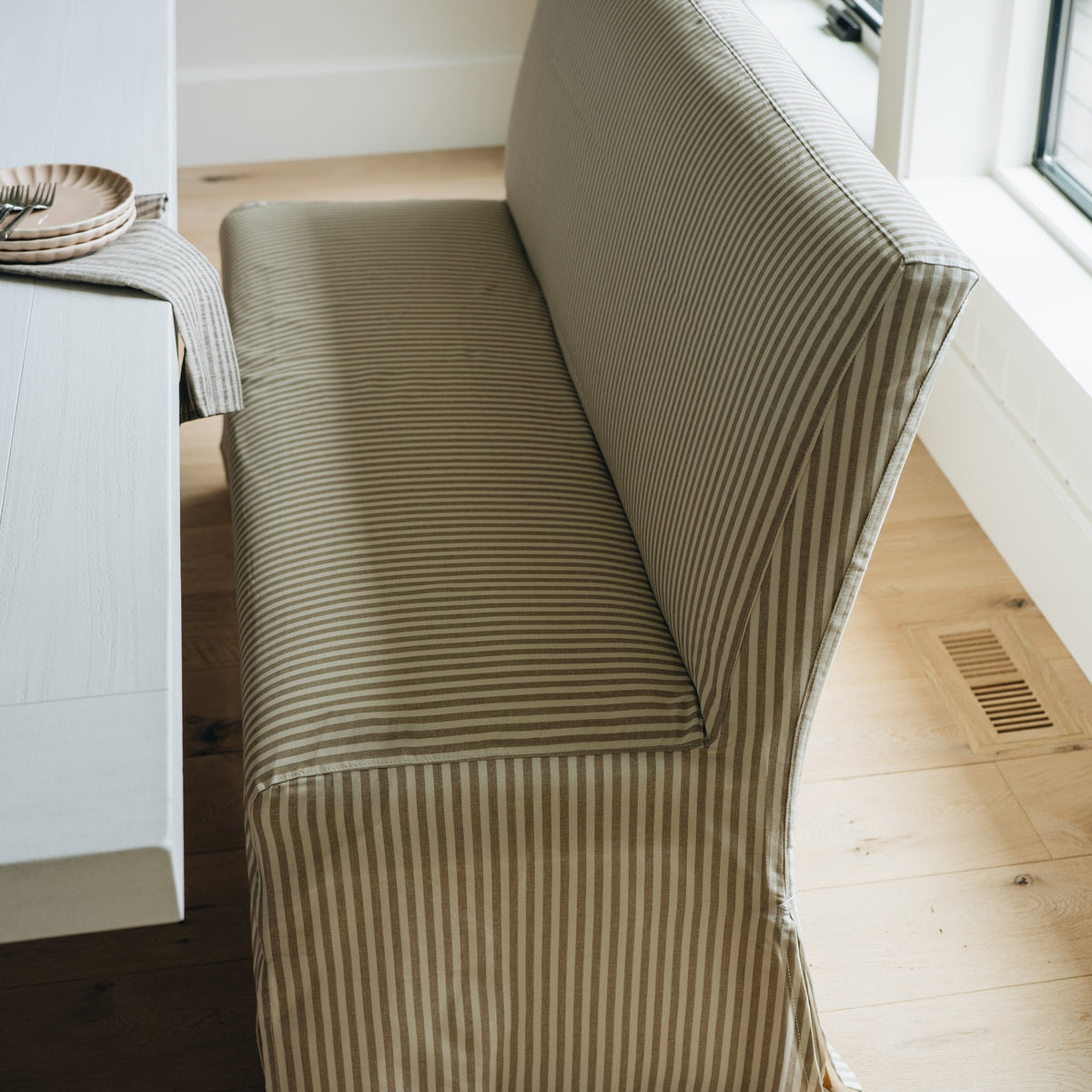 A striped upholstered bench or banquette in a warm beige and taupe fabric, shown in a bright room with light wood floors and a white table at the side.