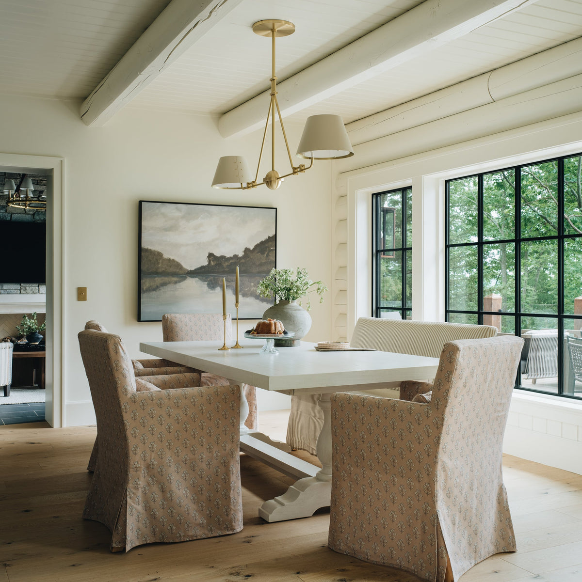 A dining room scene with a white rectangular table and four upholstered dining chairs in a light beige patterned fabric, set in a bright room with wood floors, a white paneled ceiling, and large black-framed windows.
