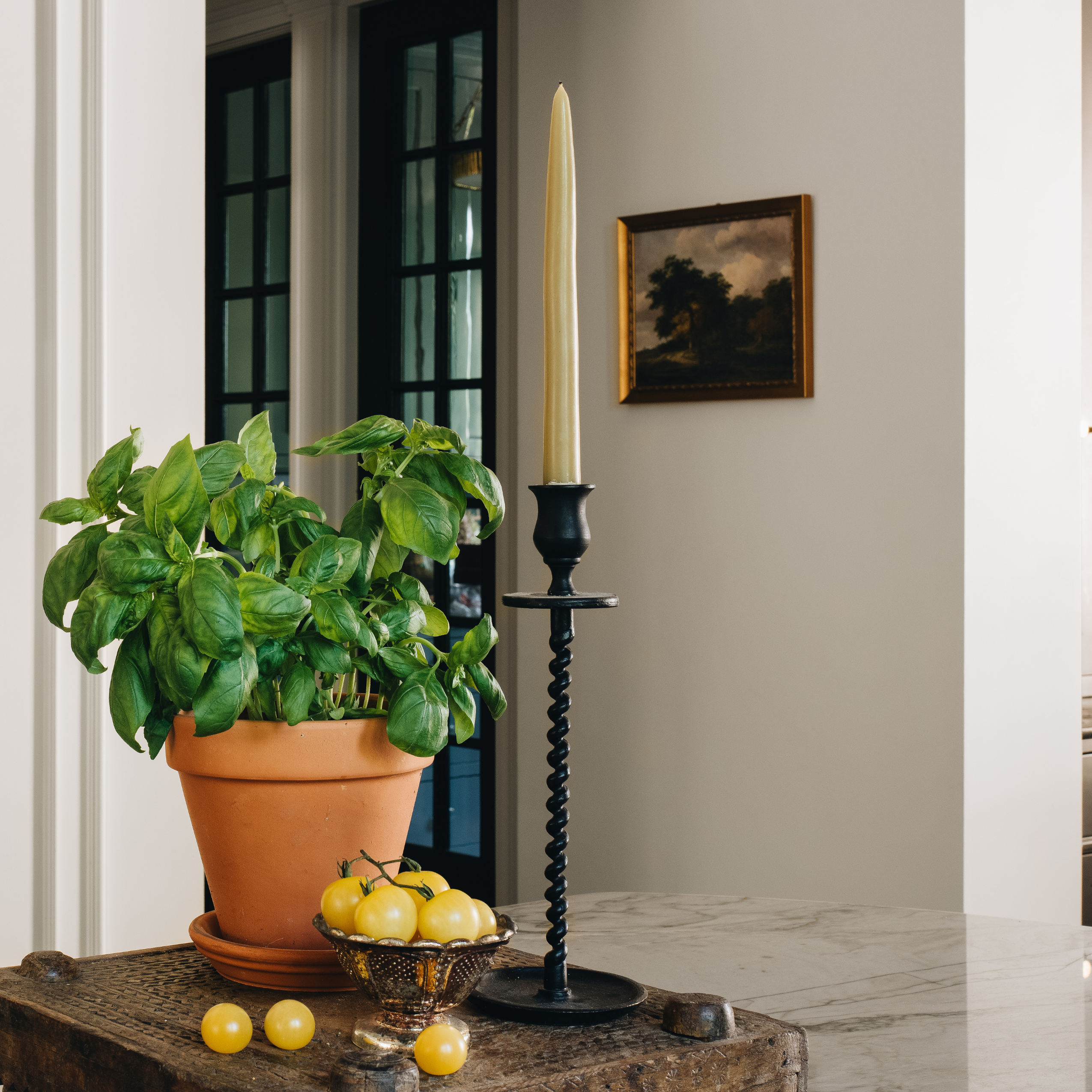 A tall black spiral candlestick holder with a cream taper candle stands on a stone surface beside a potted basil plant and lemons in a styled interior setting.
