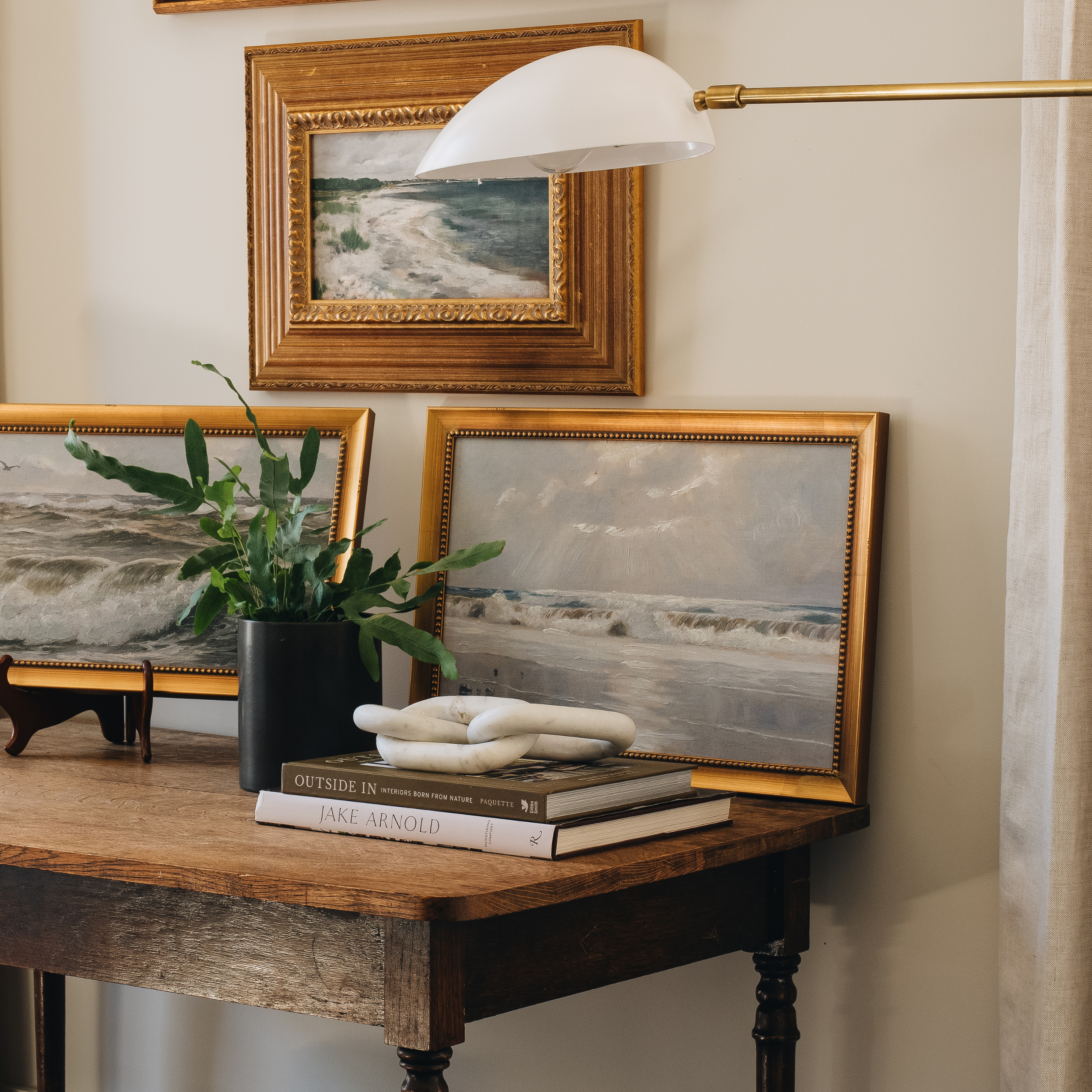A styled interior scene showing framed ocean landscape paintings on a wooden console table, with a potted plant, books, a white sculptural object, and a wall lamp above. The product appears to be the framed wall art rather than the furniture or decor props.