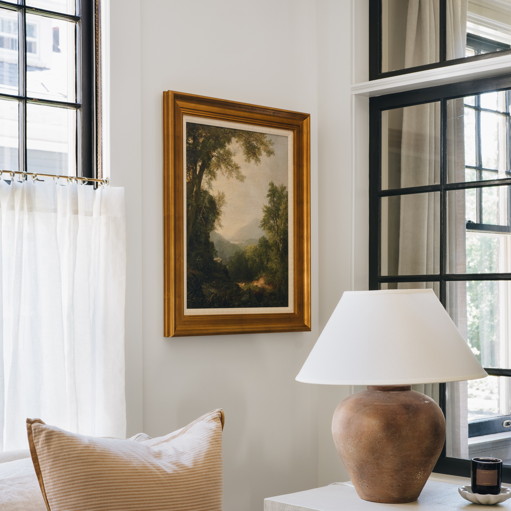 A framed landscape painting with a gold frame hangs on a white wall in a bright room, with a lamp, curtains, and windows visible.