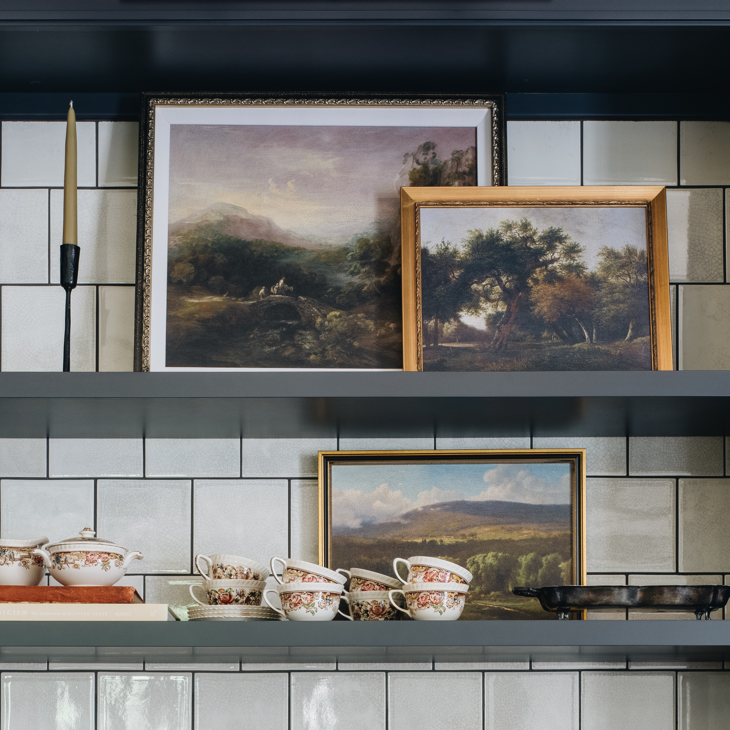 Three framed landscape paintings in different wood frames are displayed on dark gray shelves against a white subway tile wall, alongside decorative teacups and a candlestick.