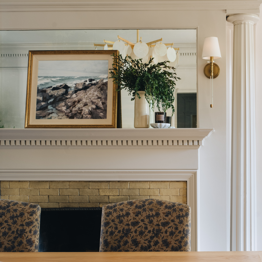 A framed landscape photograph of rocky shores in a gold frame, displayed on a white fireplace mantel alongside a vase of greenery and a wall sconce, with patterned chairs in front of a brick fireplace.