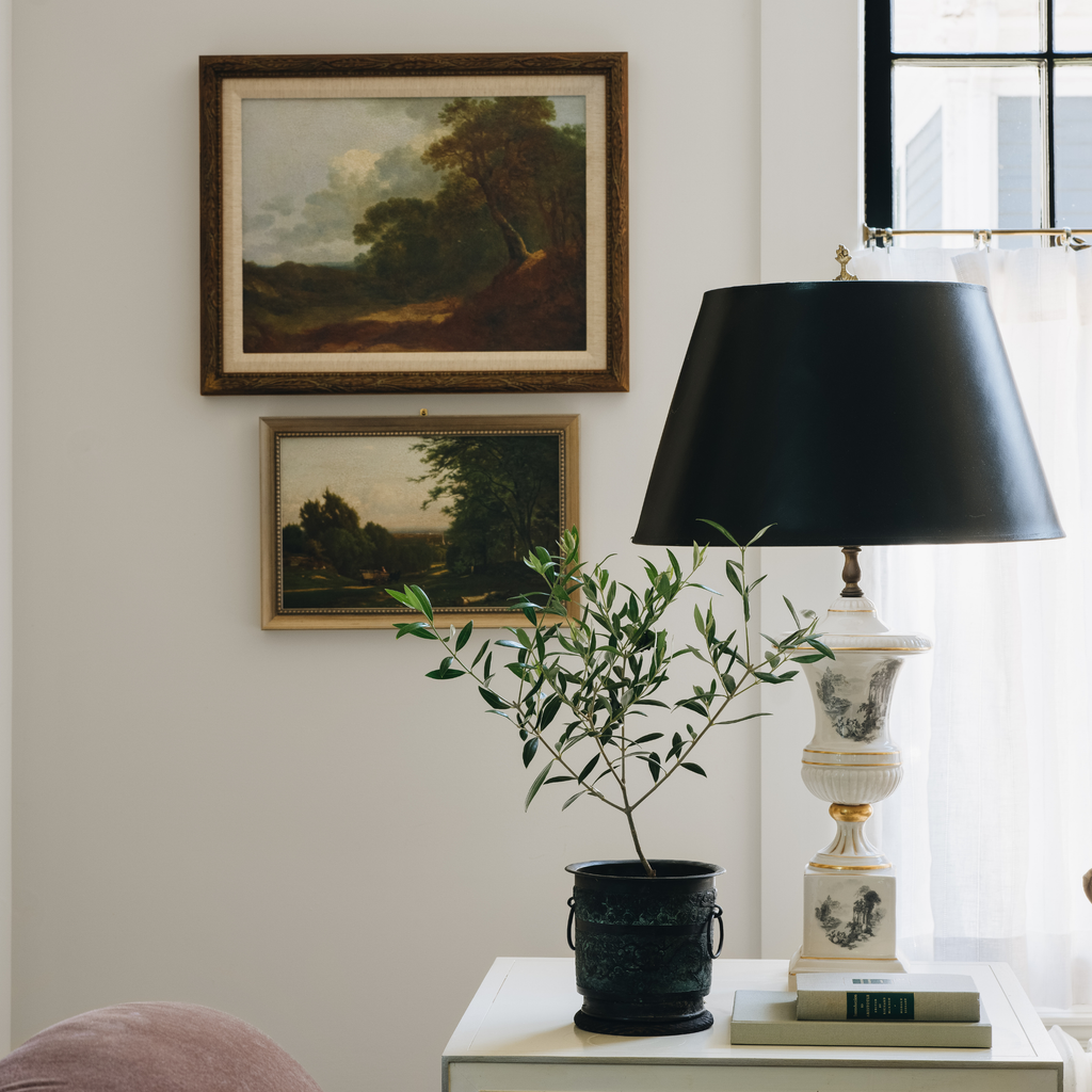 A styled interior scene with two framed landscape paintings on a light wall, a black lamp on a white table, a potted olive branch, and part of a pink upholstered chair visible in a bright room.