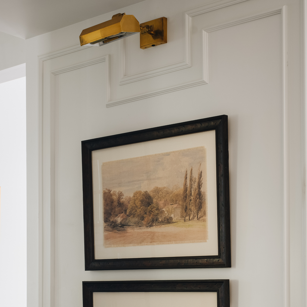 A framed landscape art print with a black frame and white mat, hanging on a white paneled wall below a gold wall sconce.