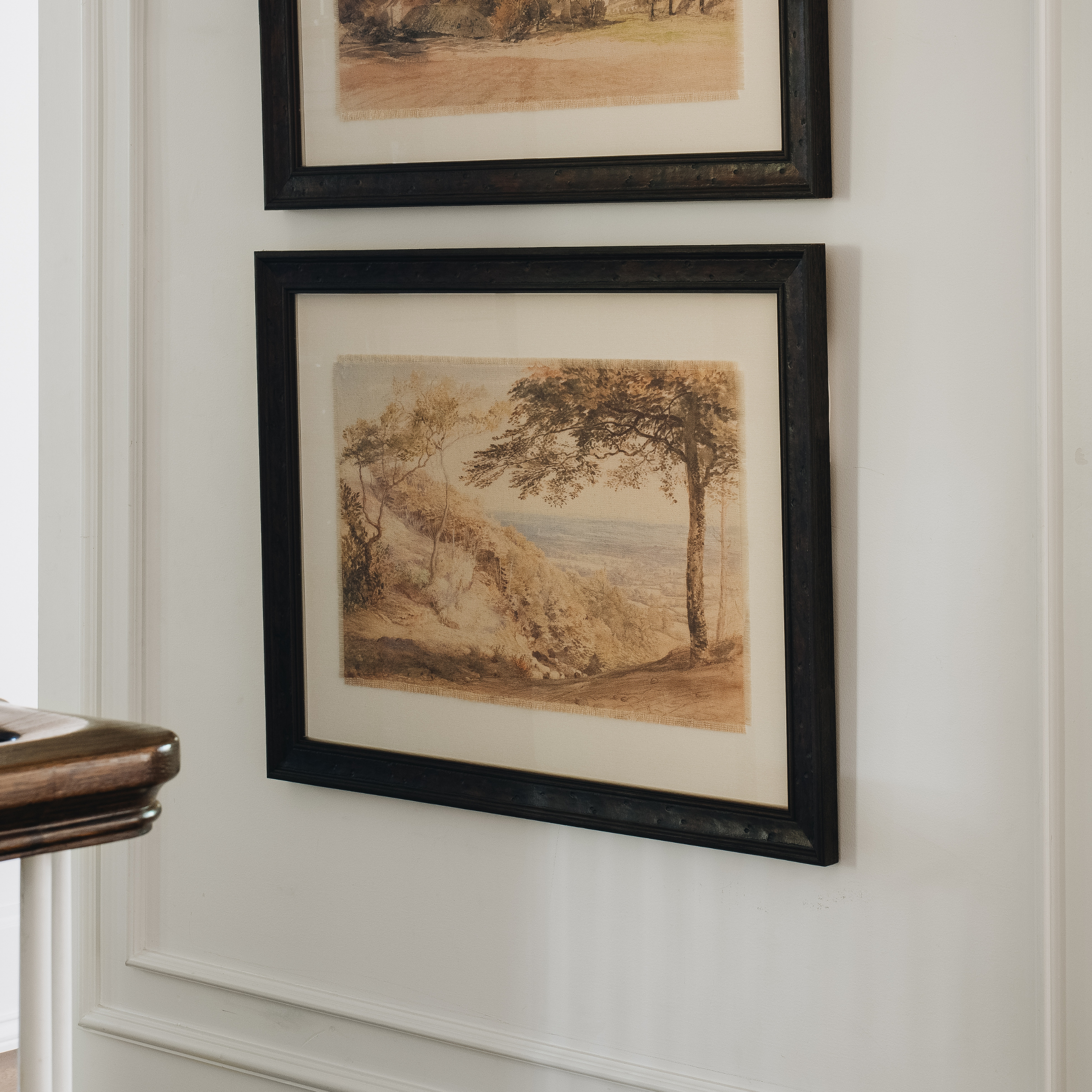 A framed landscape art print with a black frame and cream mat, hanging on a white paneled wall next to a wooden table edge.