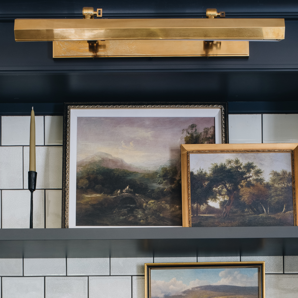 A brass wall sconce light fixture mounted on a dark navy blue shelf above framed landscape paintings and a candle holder against a white subway tile wall.