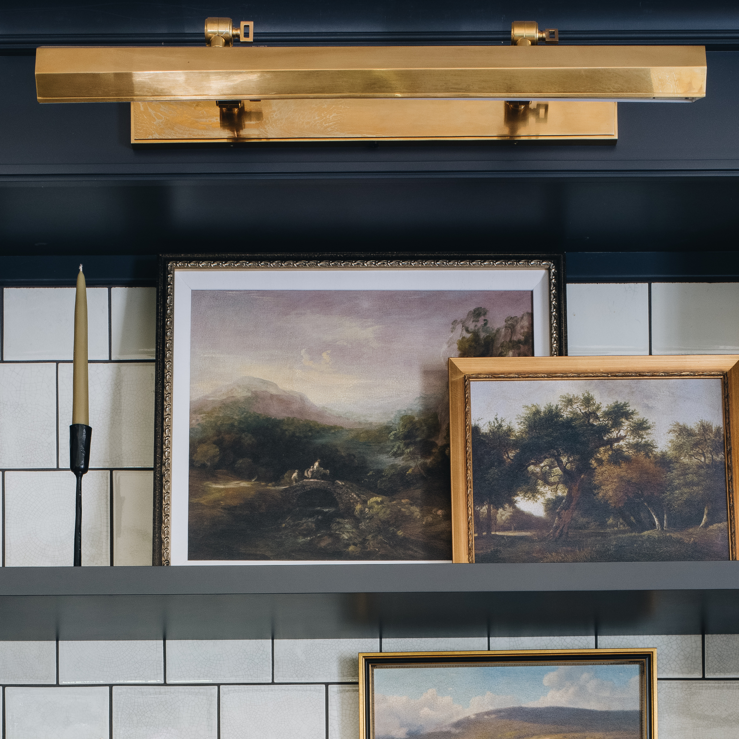 A brass wall sconce light fixture mounted on a dark navy blue shelf above framed landscape paintings and a candle holder against a white subway tile wall.