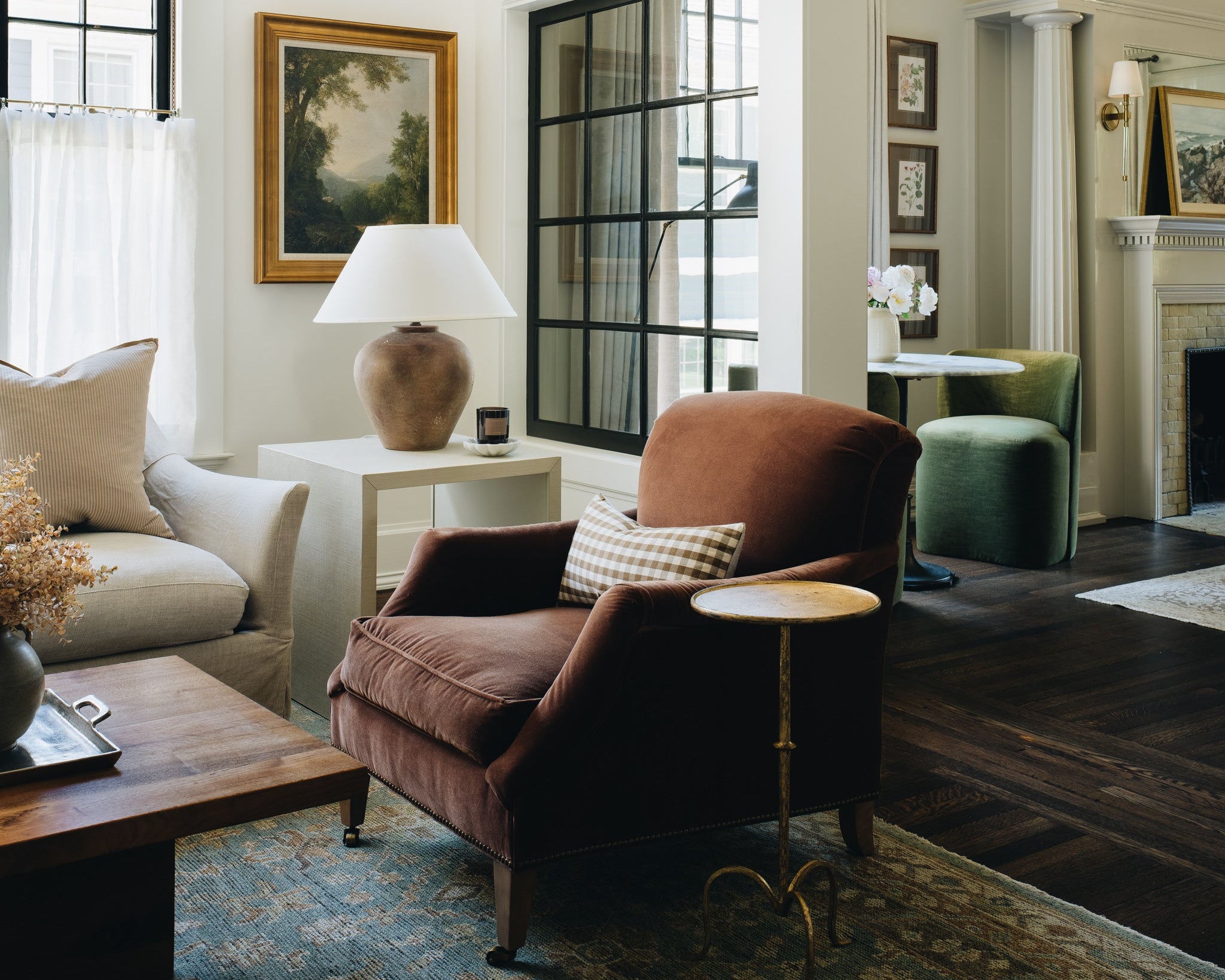 A styled interior living room scene featuring a brown velvet armchair as the central product, surrounded by a beige sofa, a side table with a lamp, a patterned rug, dark wood flooring, and framed wall art.