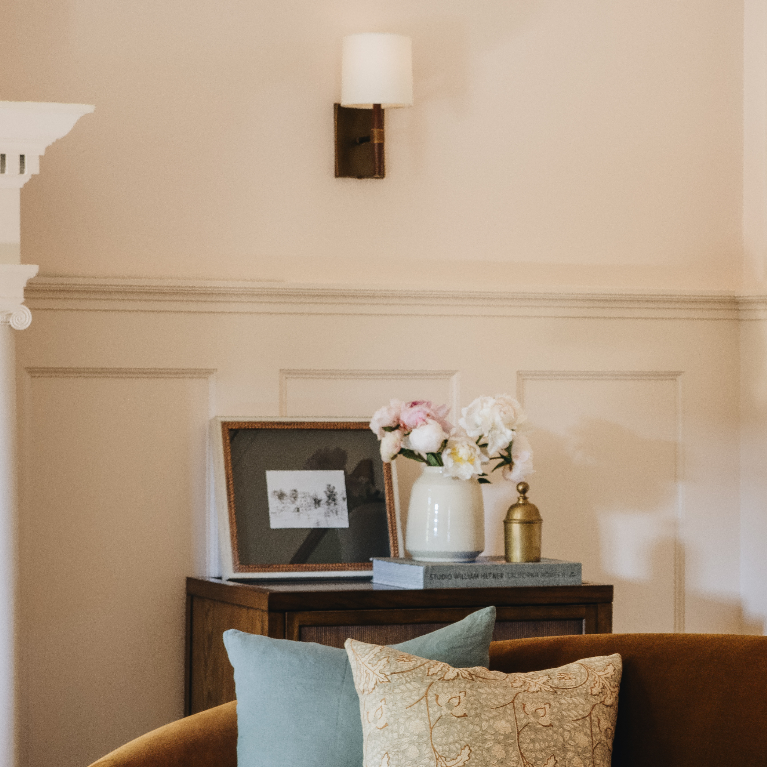 A warm beige wall with wainscoting and a wall-mounted sconce, featuring a dark wood console table with a framed print, a white vase of flowers, books, and a brass bottle. A brown velvet sofa with decorative pillows is visible in the foreground.