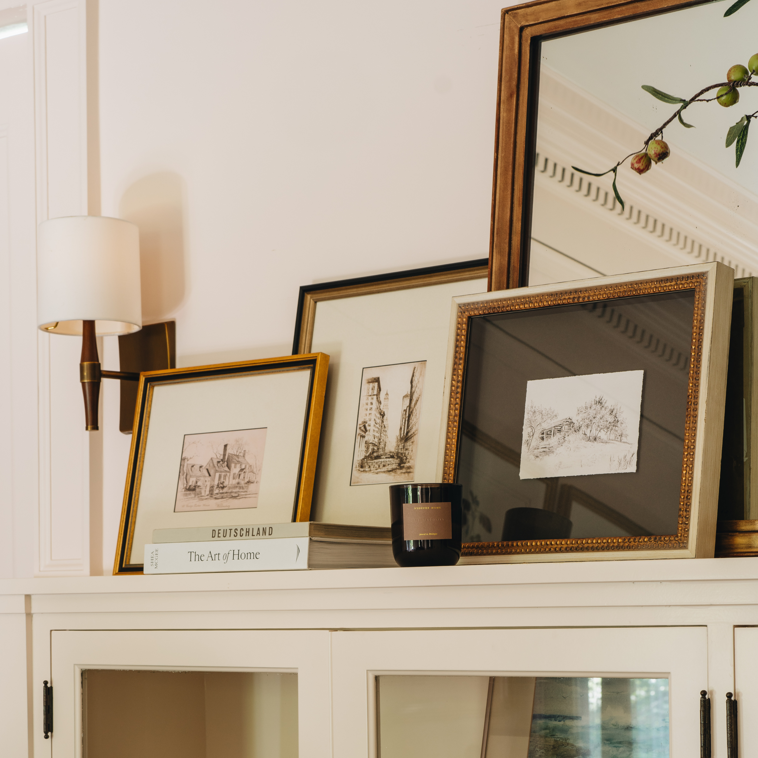 A styled interior scene showing several framed art prints and sketches displayed on a white cabinet, with a wall lamp, books, and a mirror in the background.