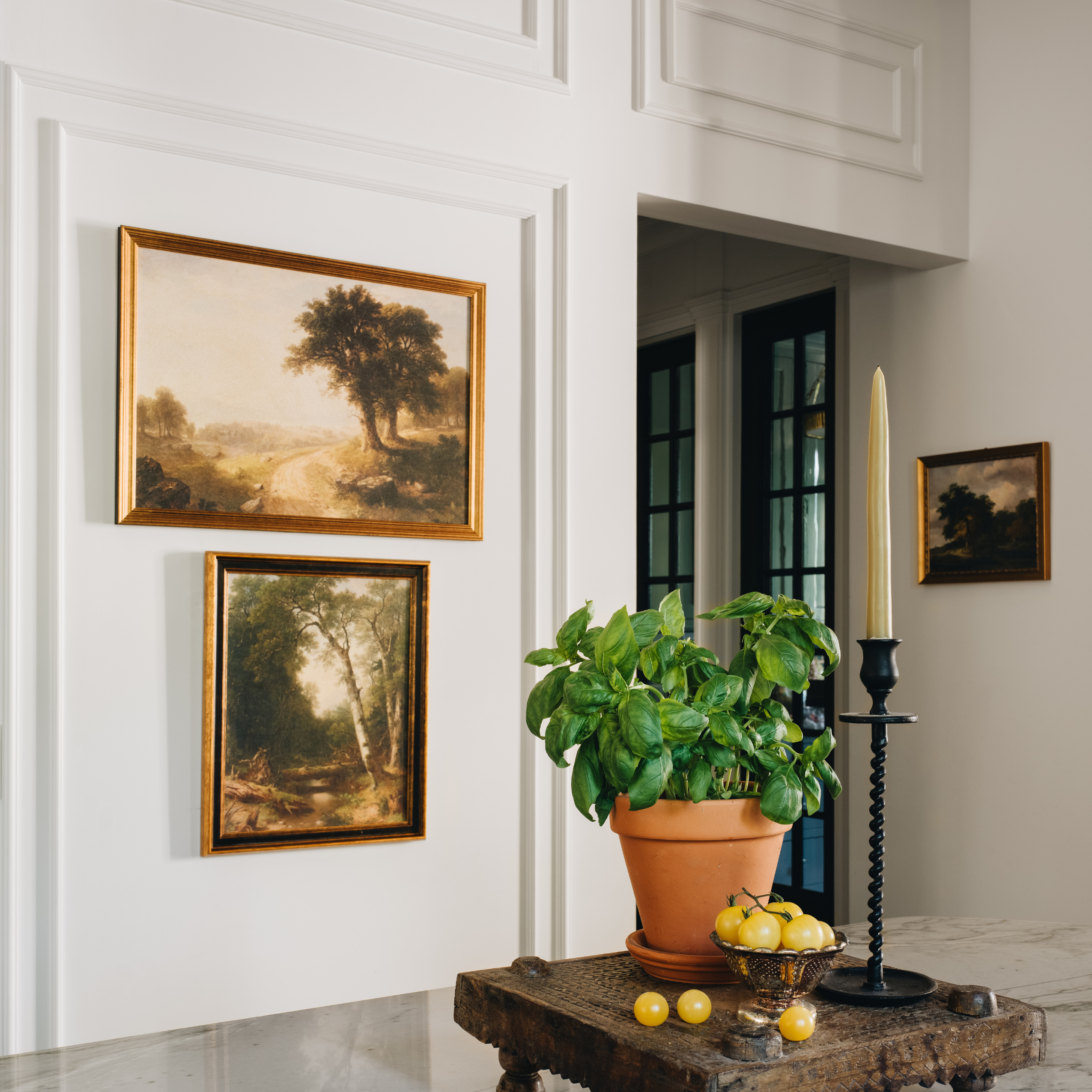 A styled interior scene showing two framed landscape paintings hanging on a white paneled wall above a rustic table with a potted basil plant, lemons, and a tall candlestick.