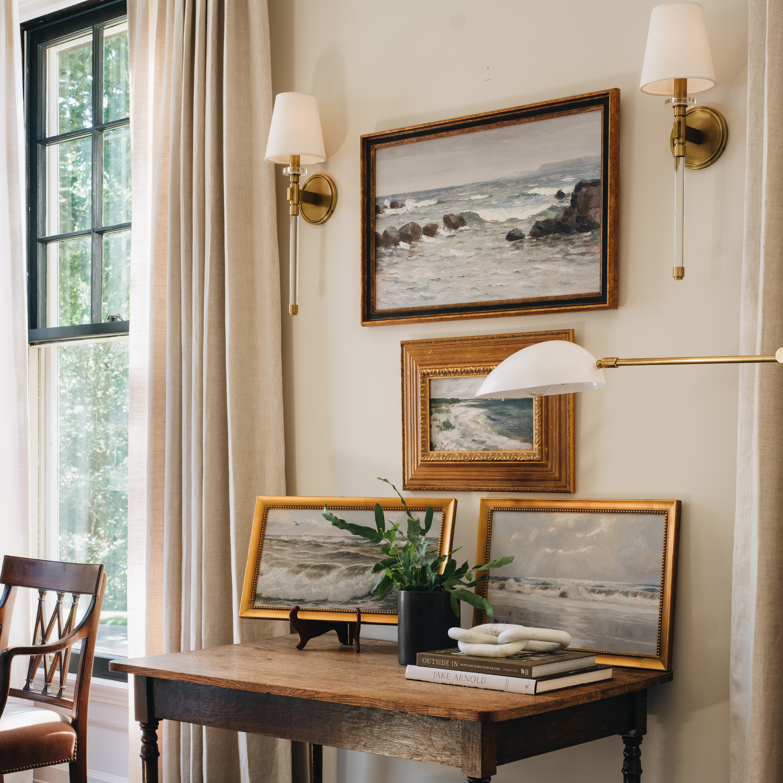 A styled interior room scene showing framed ocean landscape paintings hanging on a beige wall above a wooden desk, with wall sconces, curtains, a window, and a plant.