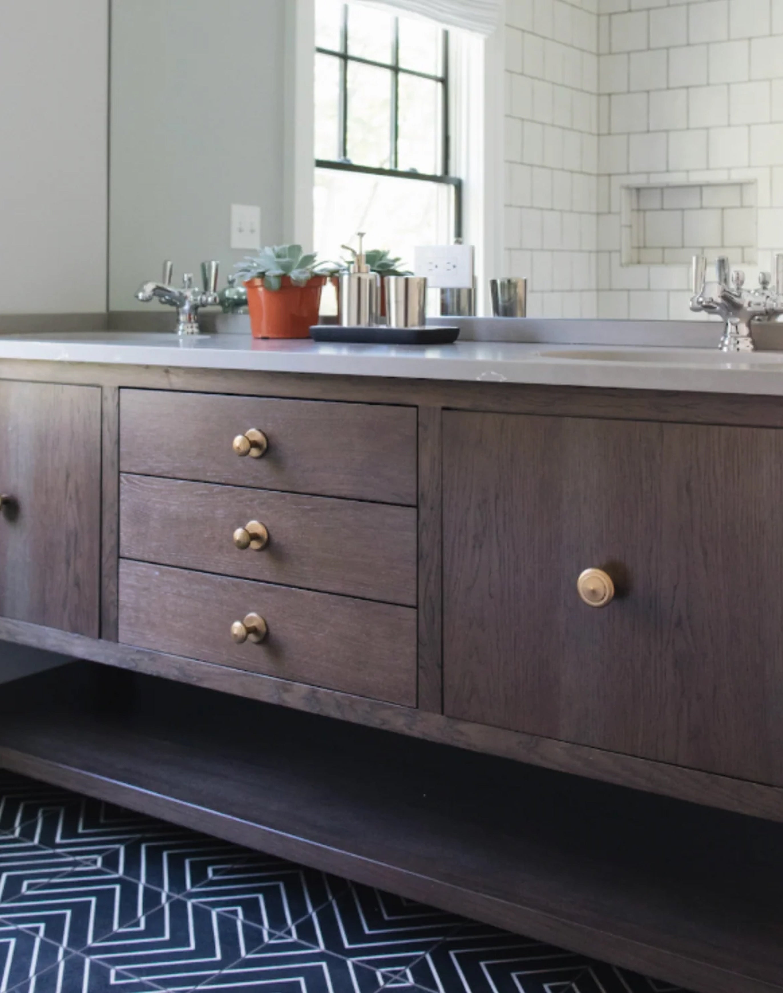 A dark brown wooden bathroom vanity with three drawers and a cabinet door, featuring brass round knobs, set against a white subway tile wall with a mirror and sink visible.