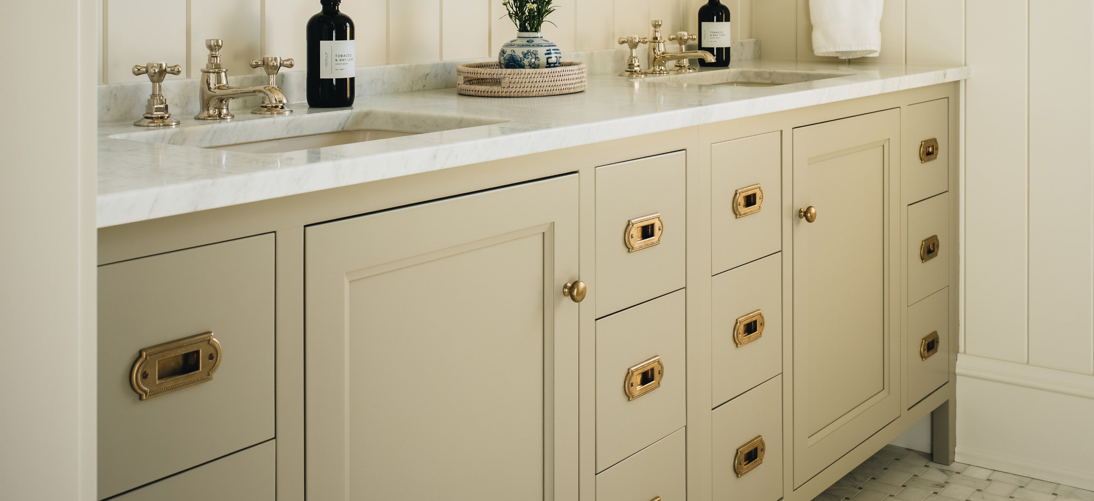 A cream-colored bathroom vanity cabinet with brass hardware, a white marble countertop, and two sinks with gold faucets, shown in a clean interior setting.