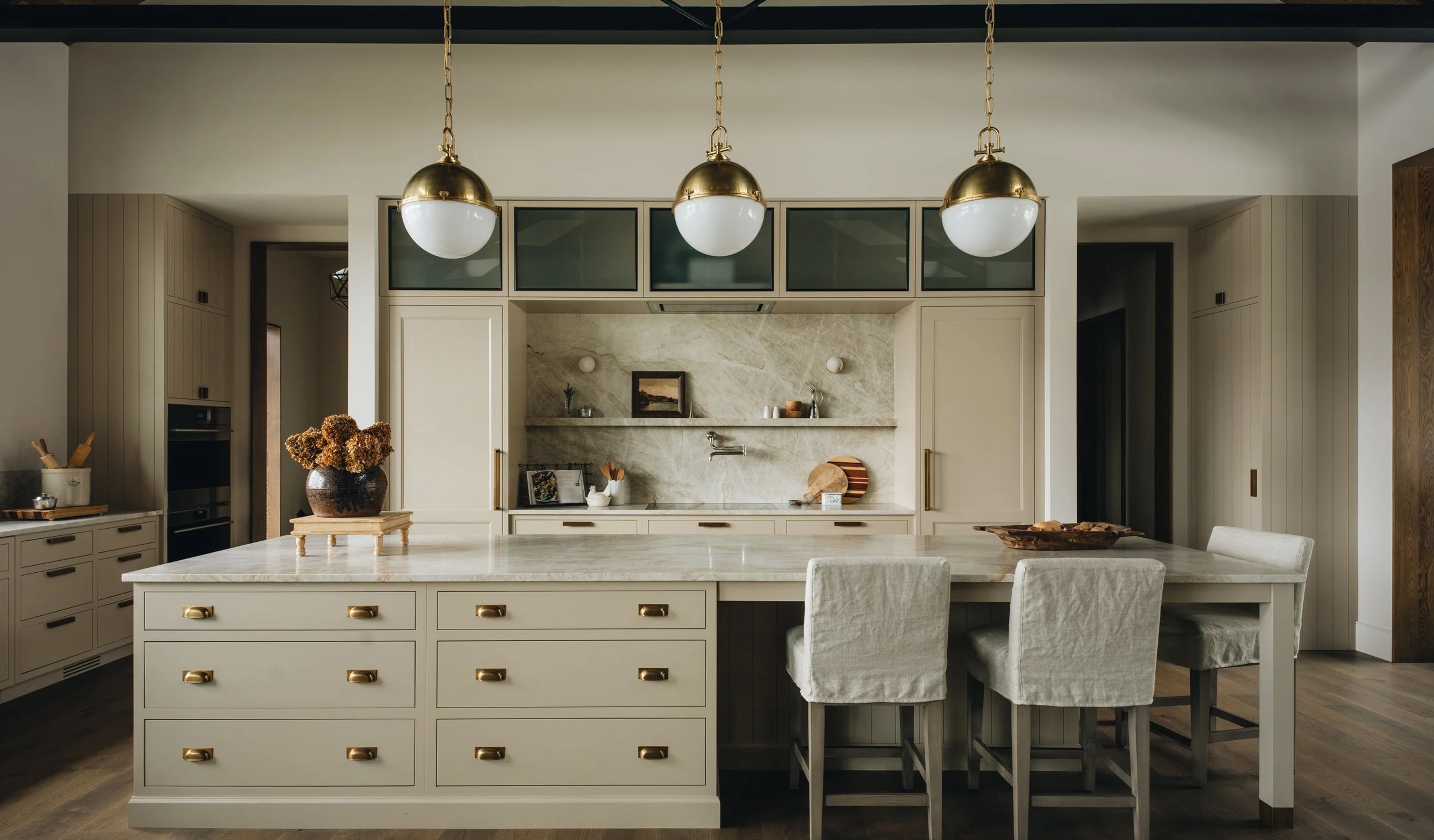 A modern kitchen island with cream-colored cabinets, marble countertops, brass hardware, and three hanging pendant lights above. Two white bar stools are tucked under the counter, and the room has warm wood flooring and neutral walls.