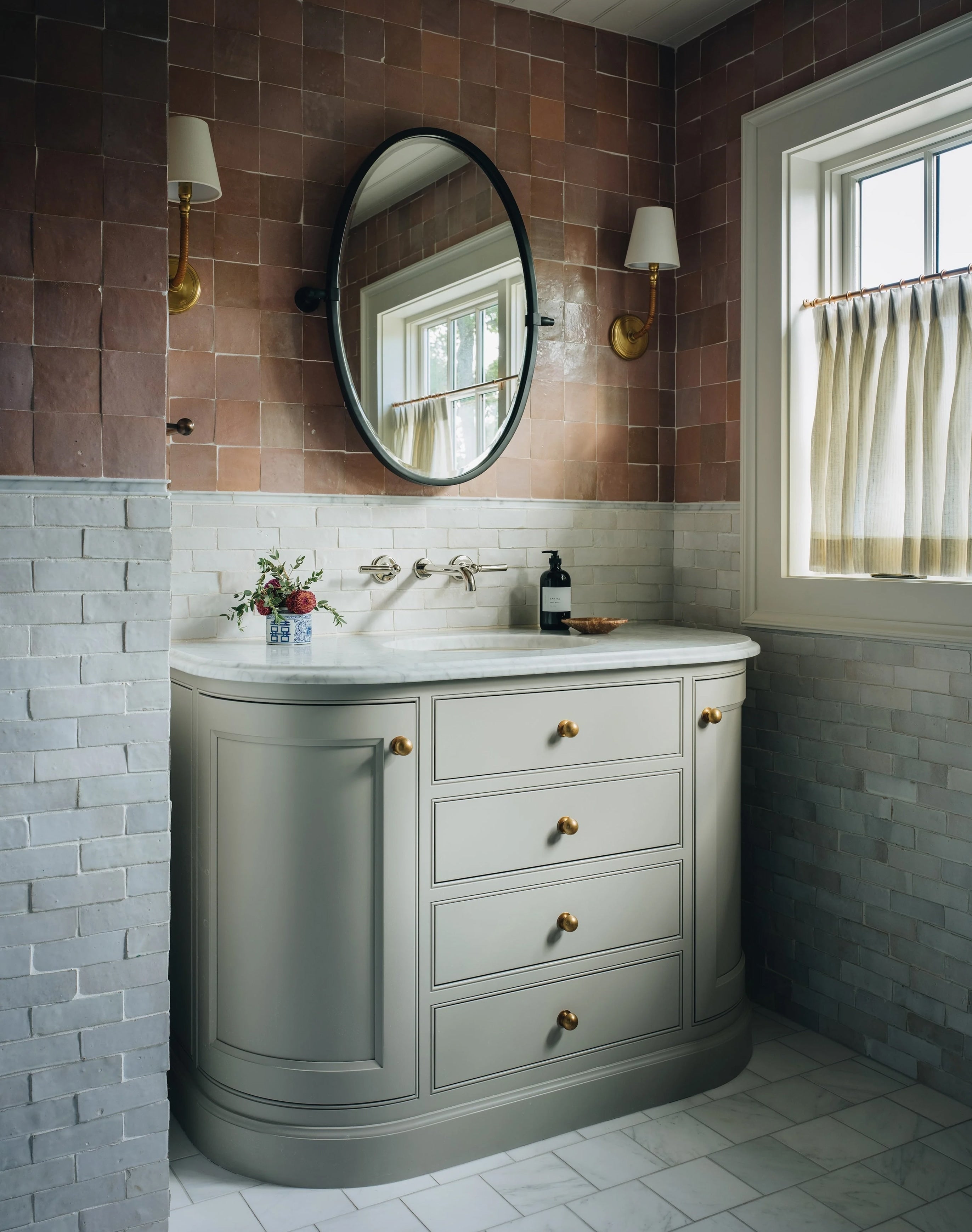 A classic curved bathroom vanity cabinet in a muted gray-green, featuring brass knobs, a white countertop with an integrated sink, and a round black-framed mirror. The vanity is set against a rustic wall with a brown upper section and a light gray lower section, flanked by two gold wall sconces.