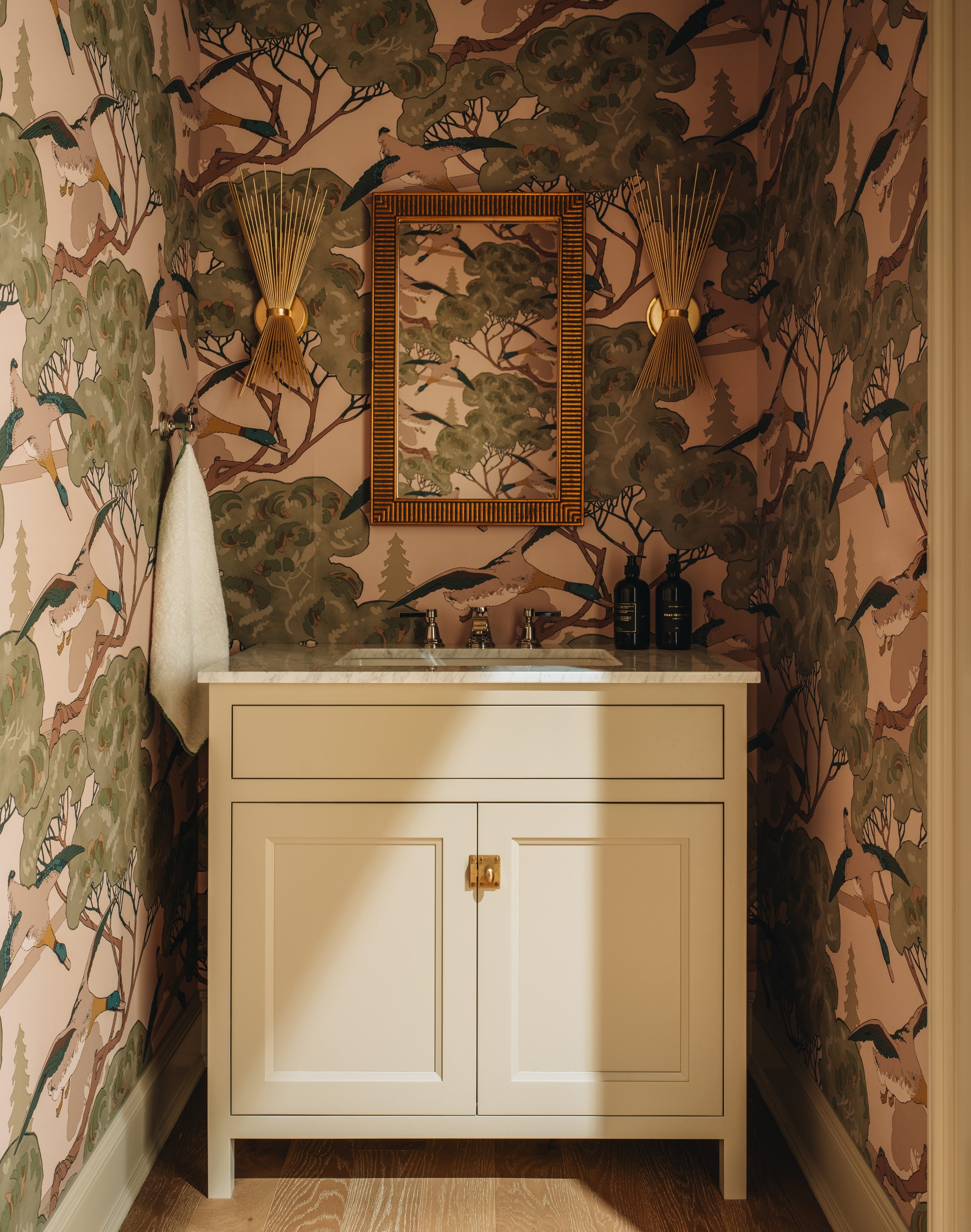 A bathroom vanity with a cream-colored cabinet and white marble countertop, set against wallpaper featuring a detailed forest and bird pattern in earthy tones. A matching decorative mirror with a gold frame hangs on the wall, flanked by two gold sconces.