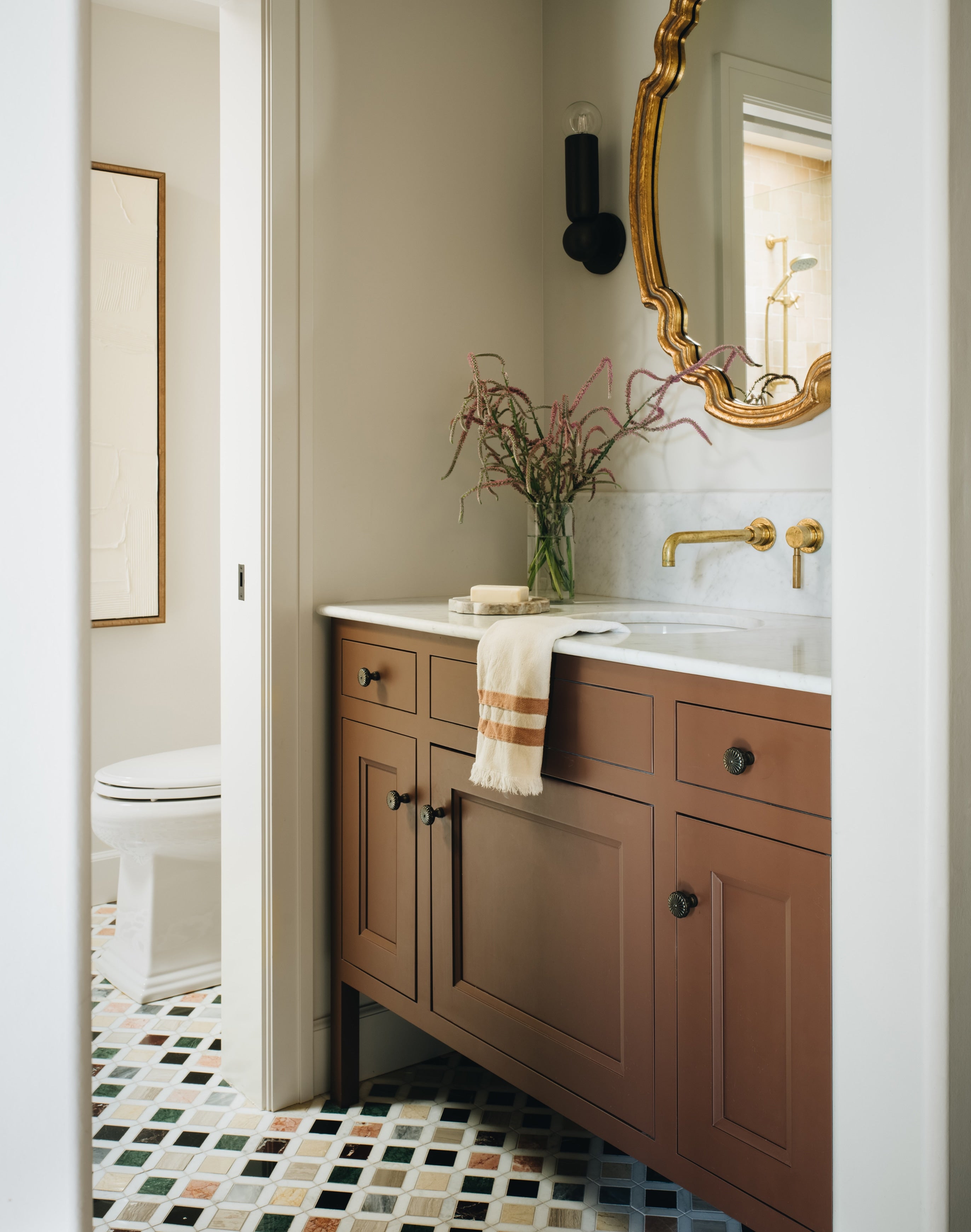 A bathroom vanity with medium brown paneled cabinets, a white countertop, a gold faucet, and a large ornate gold mirror. A white towel with tan stripes hangs from the cabinet, and a vase with dried branches sits on the counter.