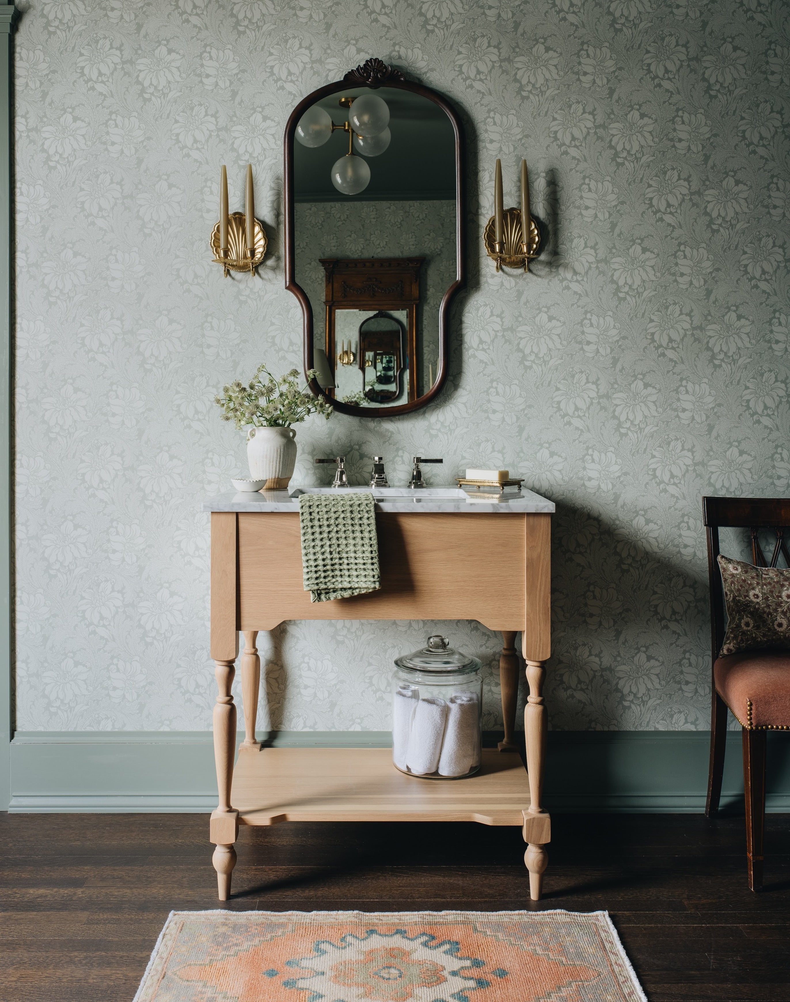 A vintage-style wooden vanity console with a white marble top and open lower shelf, styled in a room with patterned wallpaper, an ornate wall mirror, gold sconces, a towel draped over the front, and a rug on the floor.