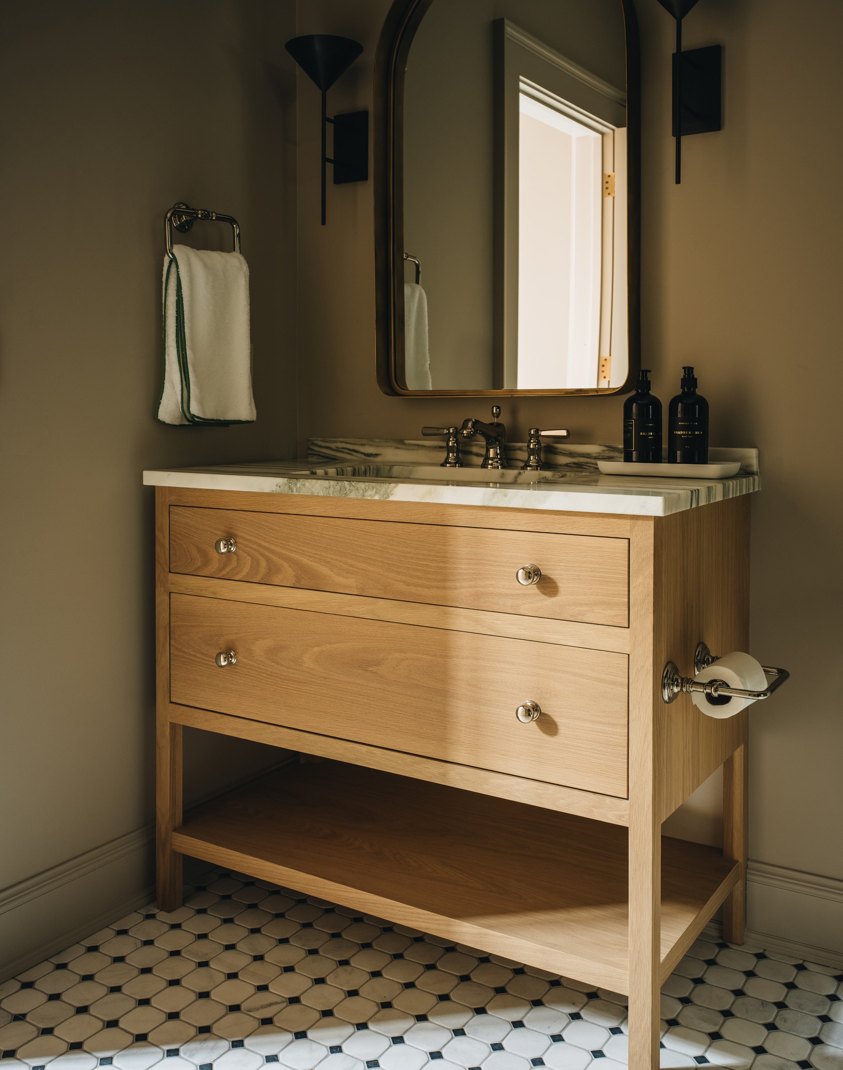 A light natural wood bathroom vanity with three drawers, an open lower shelf, and a white marble countertop, shown in a styled bathroom setting with a mirror, wall sconces, a towel, and a black-and-white patterned tile floor.
