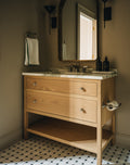 A light natural wood bathroom vanity with three drawers, an open lower shelf, and a white marble countertop, shown in a styled bathroom setting with a mirror, wall sconces, a towel, and a black-and-white patterned tile floor.