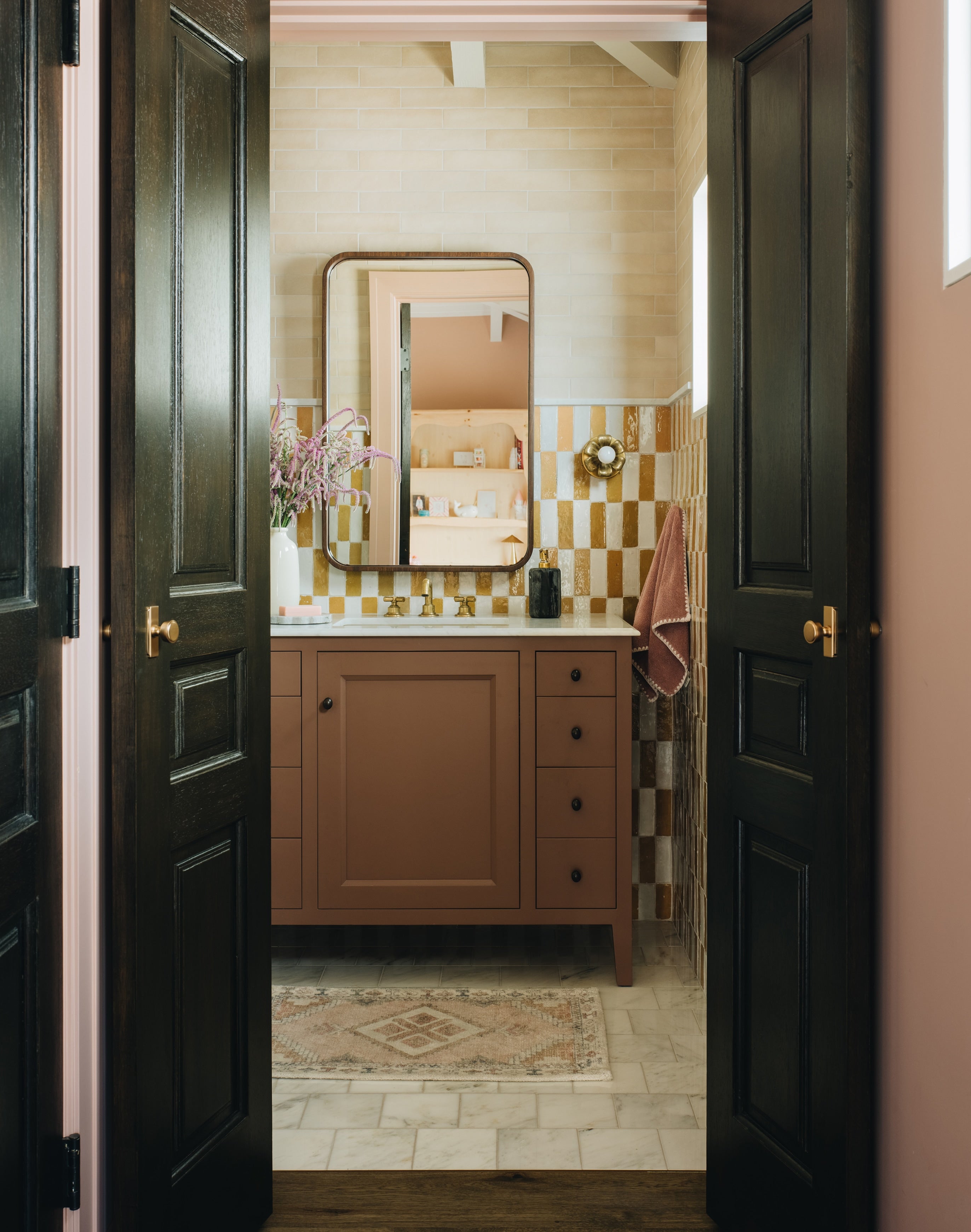A view through open dark wooden doors into a bathroom with a warm brown vanity, checkered tile backsplash, cream walls, and a patterned rug.