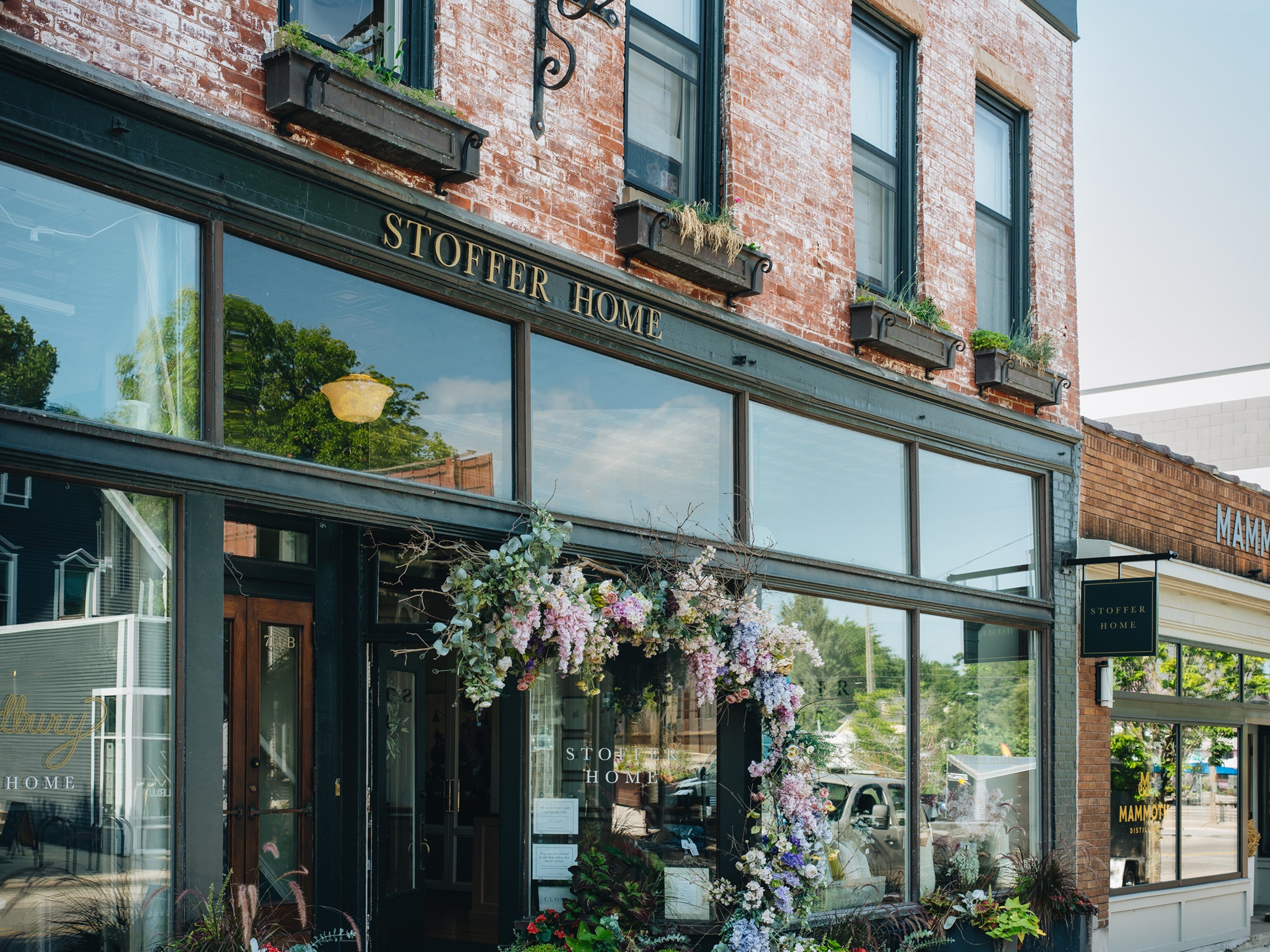 Stoffer Home storefront on a brick building with large glass windows, dark trim, hanging flower garlands, and a visible Stoffer Home sign above the entrance.