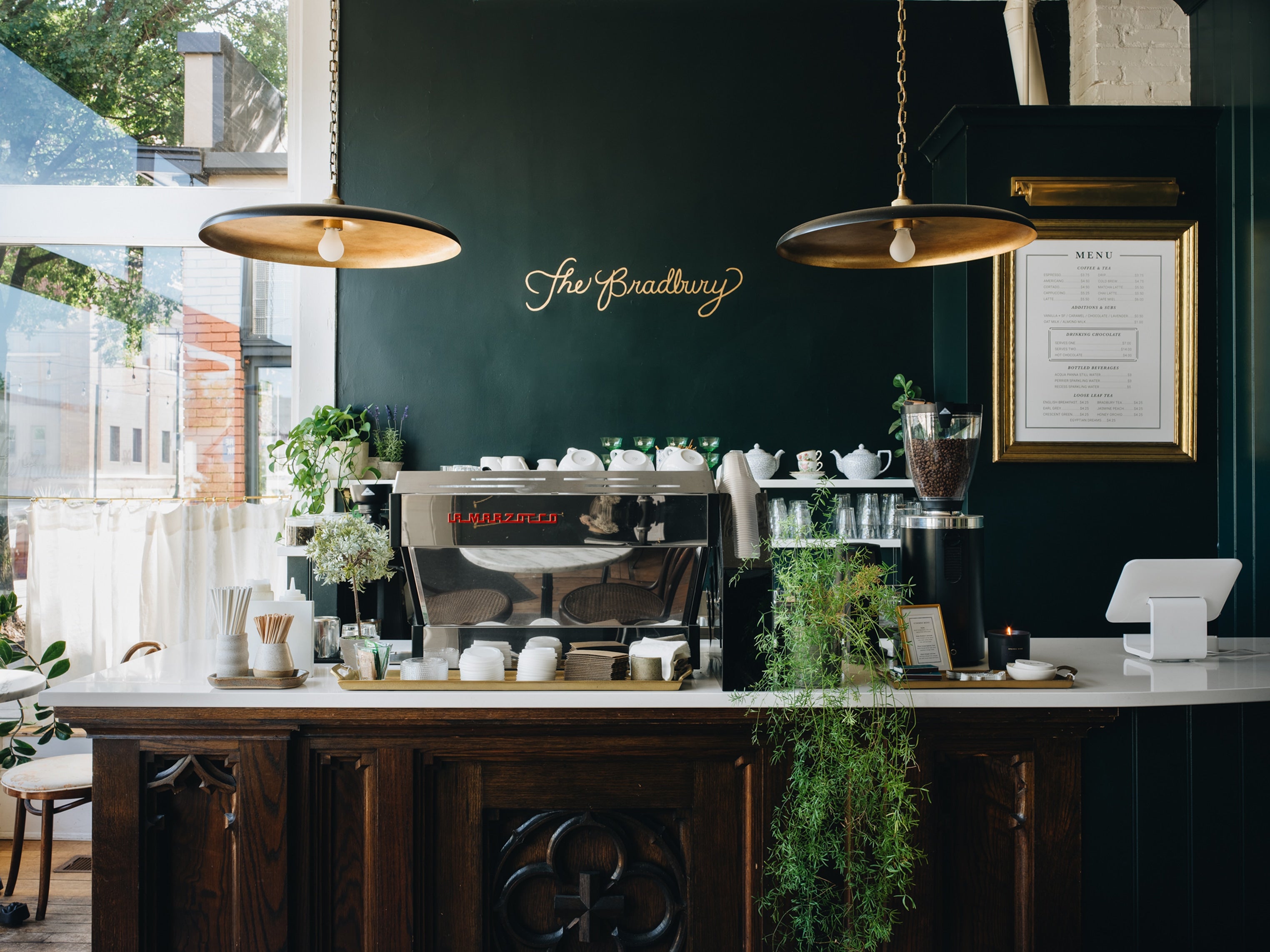 A cozy cafe interior with a dark wood counter, espresso machine, hanging pendant lights, and a dark green wall featuring the gold script logo 'The Broadway'.