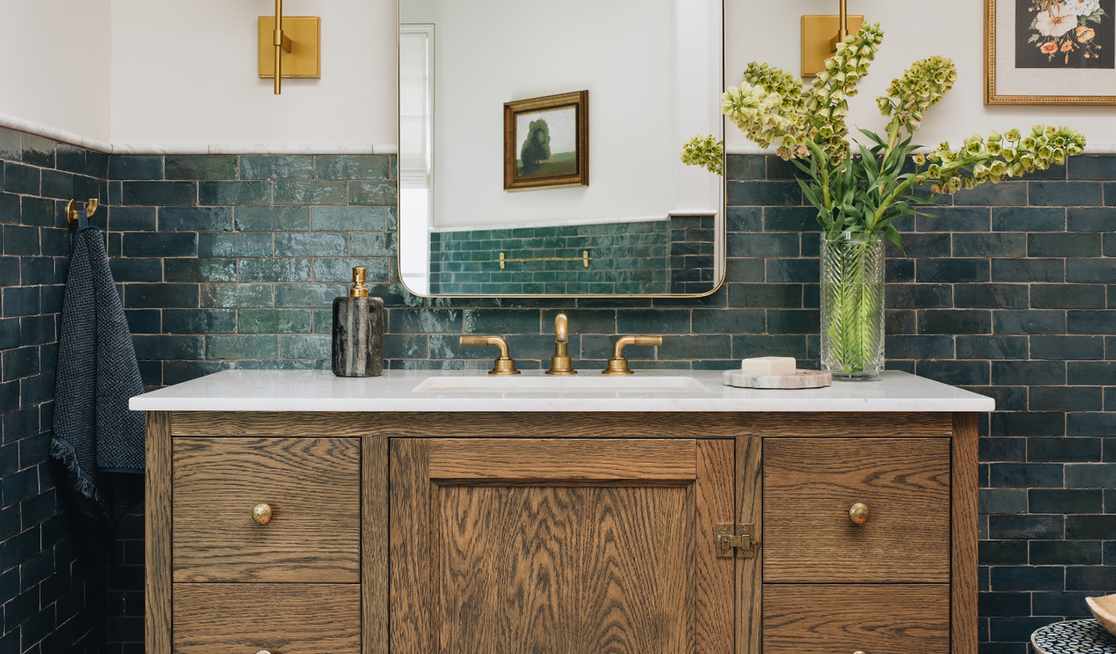 A rustic wooden bathroom vanity with a white marble countertop, integrated sink, and polished brass faucets, set against a dark green subway tile wall with a large mirror and a vase of green flowers on top.