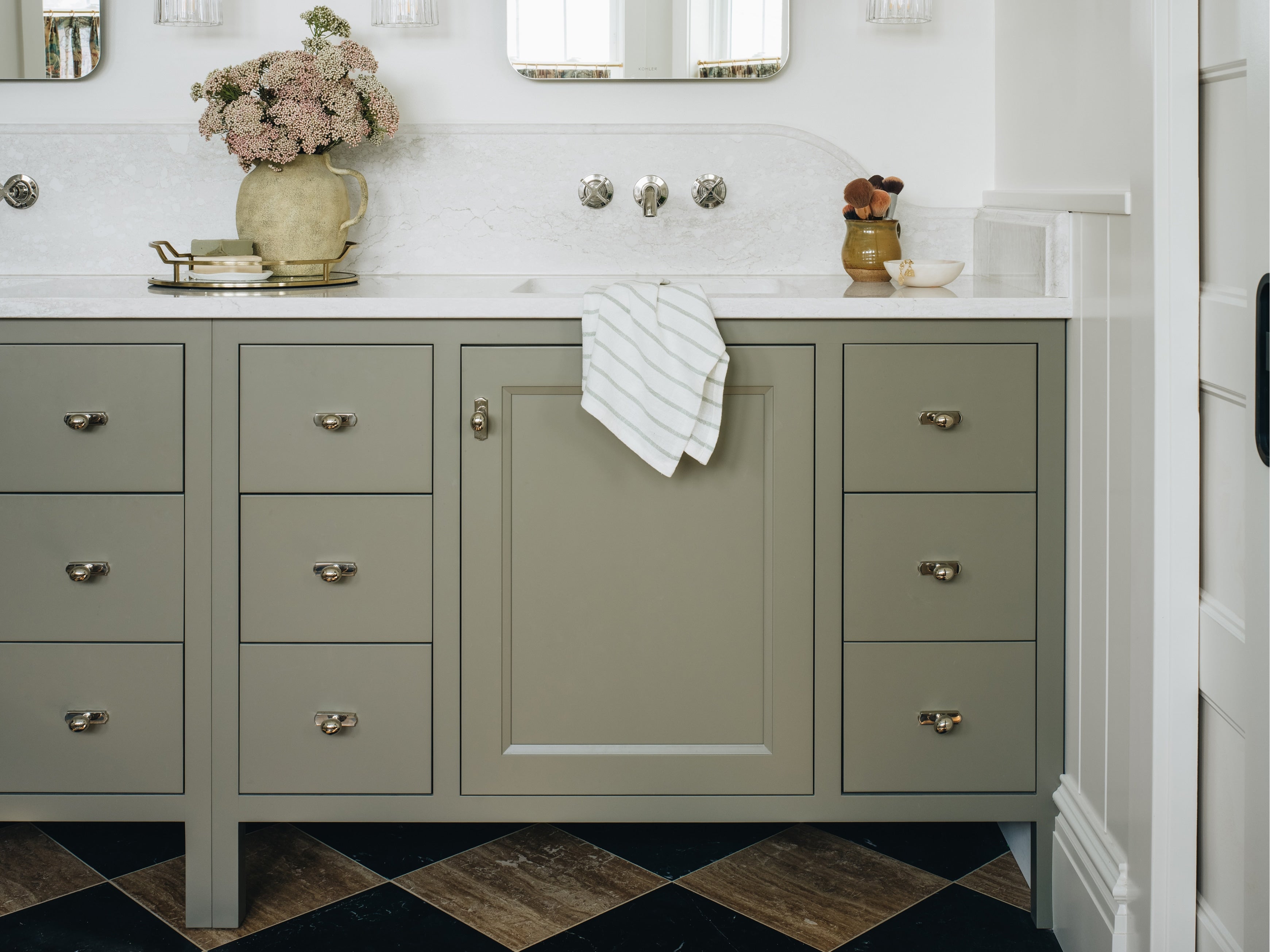 A bathroom vanity cabinet in a muted sage green color, featuring a white countertop, chrome faucets, a striped white towel, and a vase of pink flowers on a checkered dark wood and black floor.
