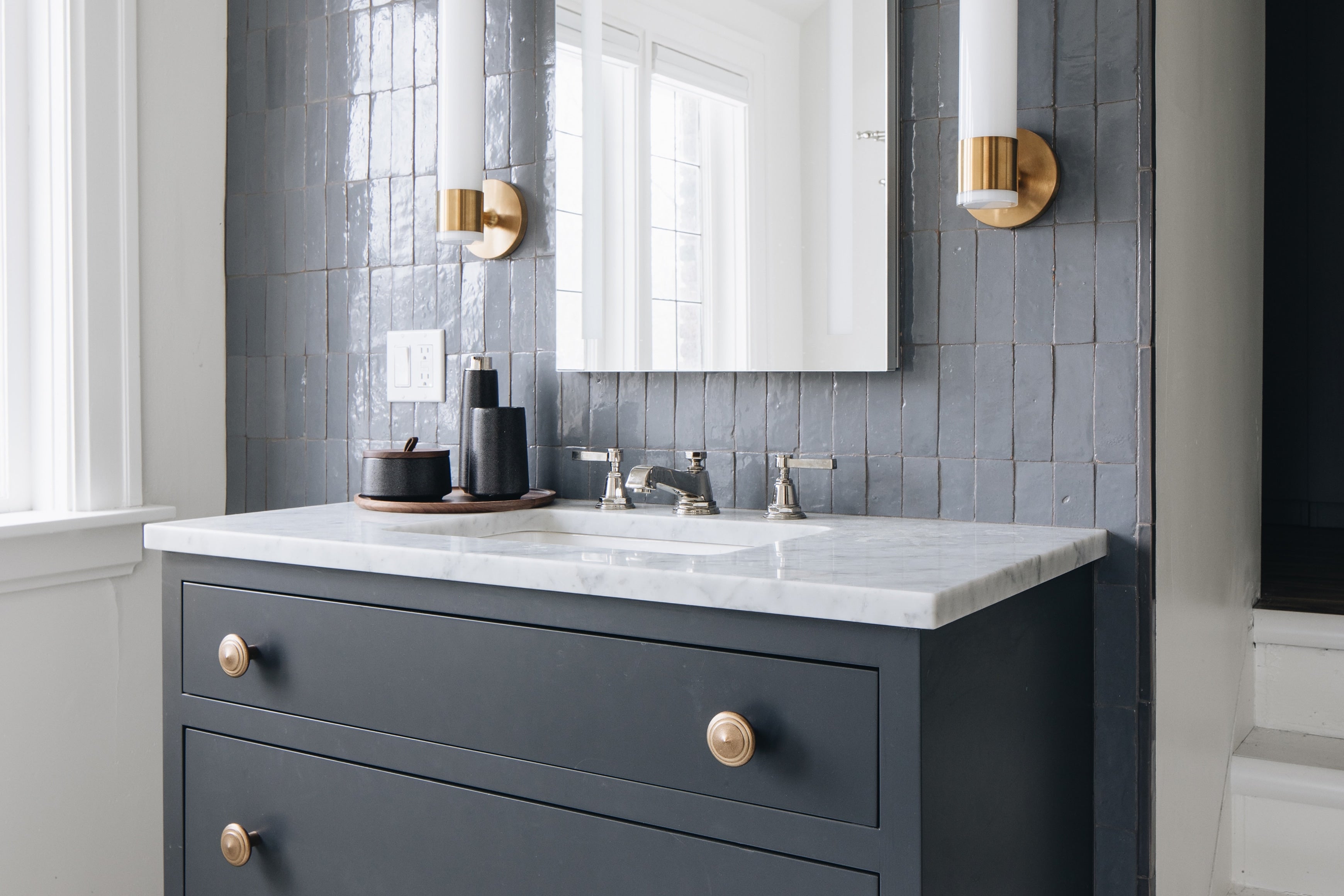 A modern bathroom vanity featuring a dark charcoal gray cabinet with brass knobs, a white marble countertop, a chrome faucet, and a gray subway tile backsplash.