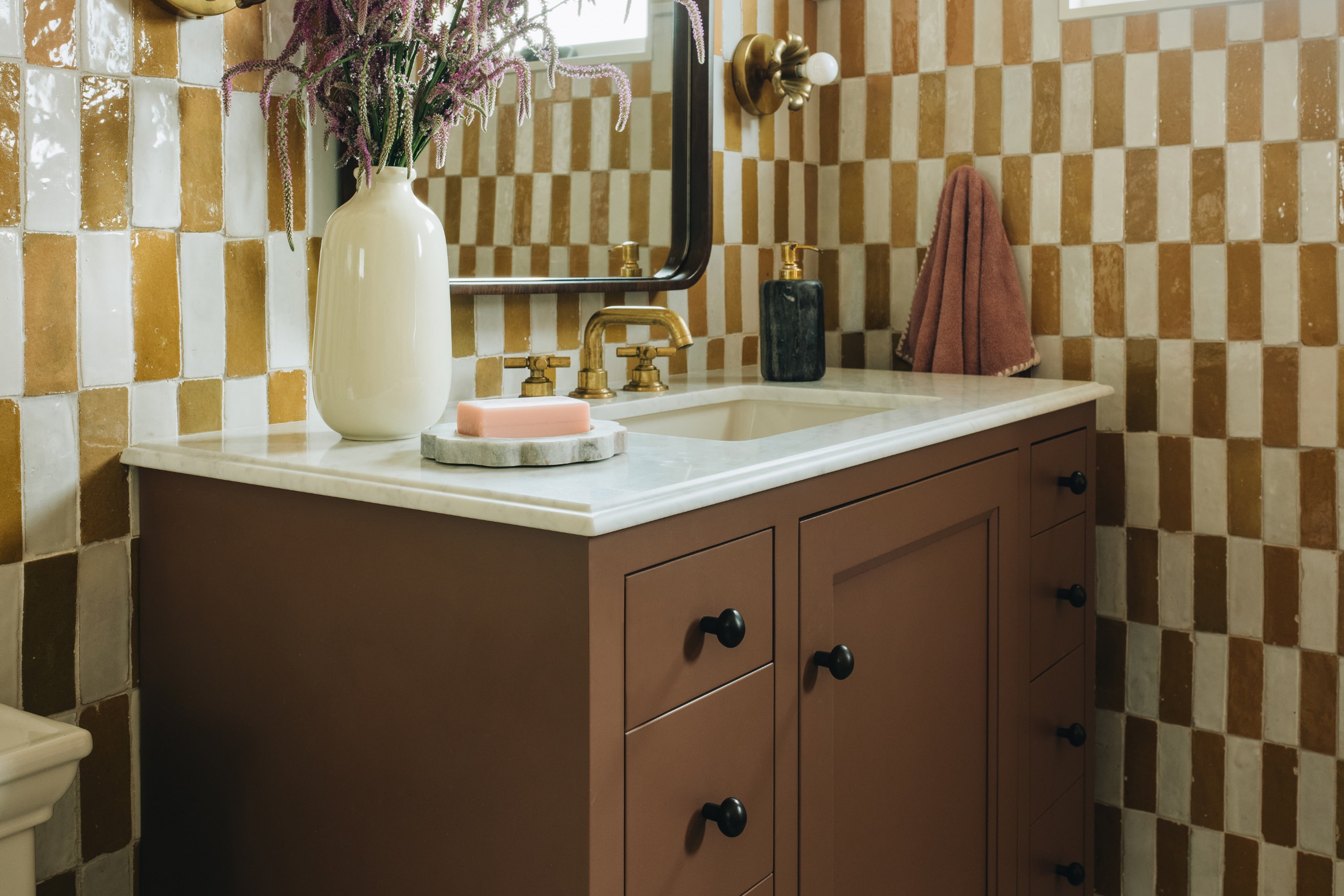 A bathroom vanity with a brown cabinet, white countertop, and a white-and-brown checkered tile wall. A large cream vase with dried flowers, a gold faucet, and a folded terracotta towel are visible.