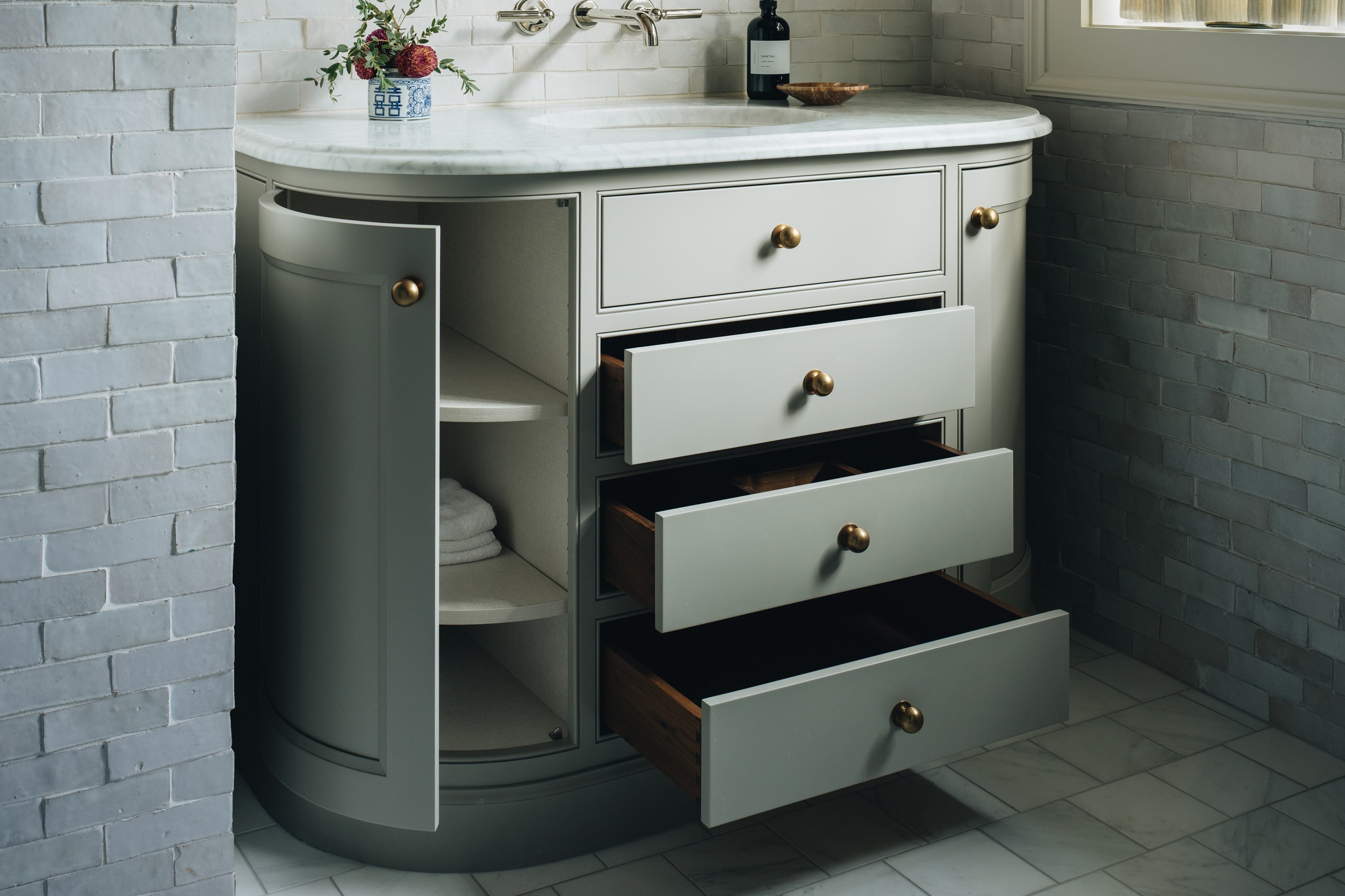 A curved bathroom vanity cabinet in a light gray finish with three open drawers showing dark wood interiors, set against a gray brick-tiled wall. A white marble countertop with an integrated sink is visible at the top, along with small decorative items.