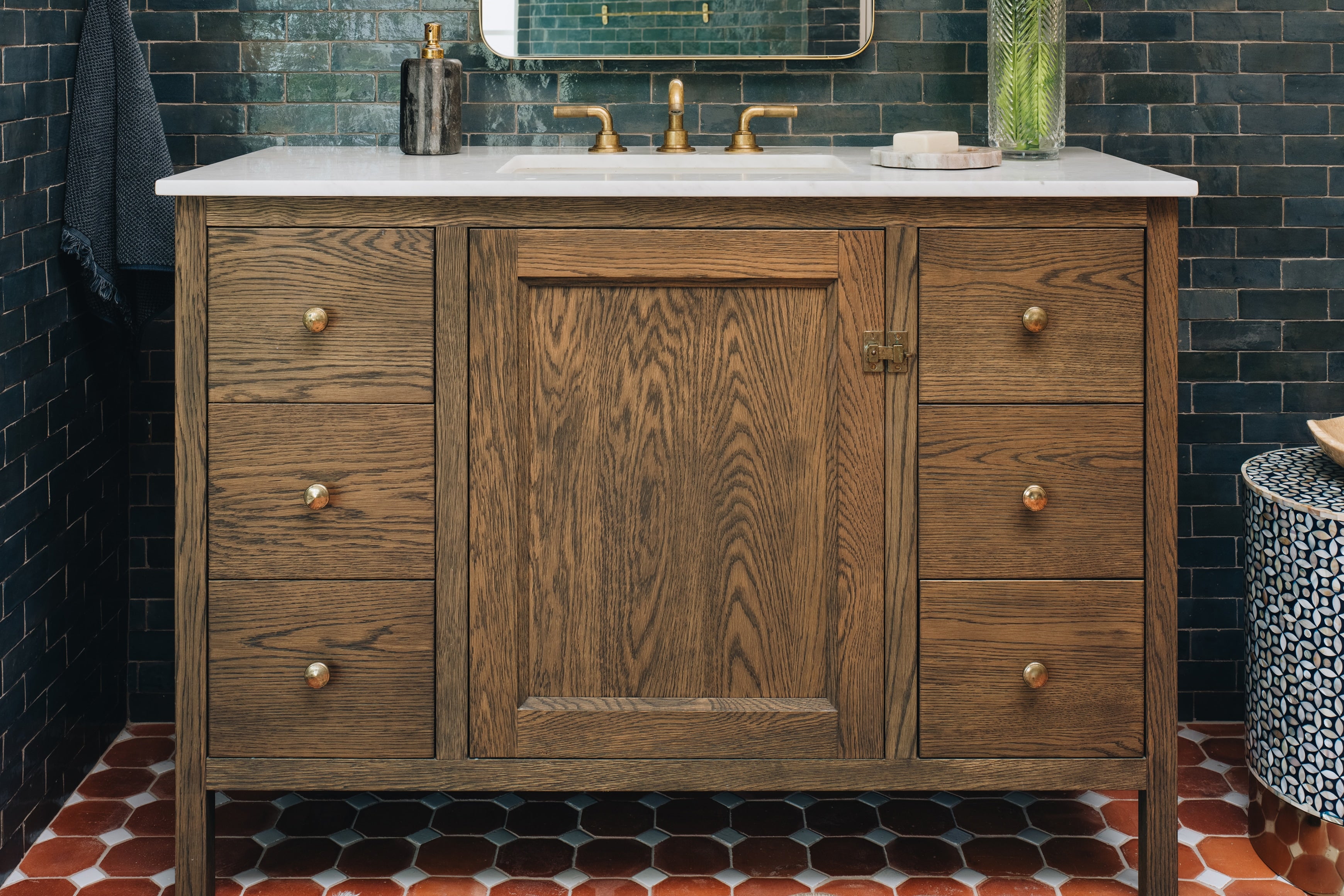 A rustic brown wooden bathroom vanity with a white marble countertop, integrated sink, and brass faucet, set against a dark tiled wall and patterned floor.